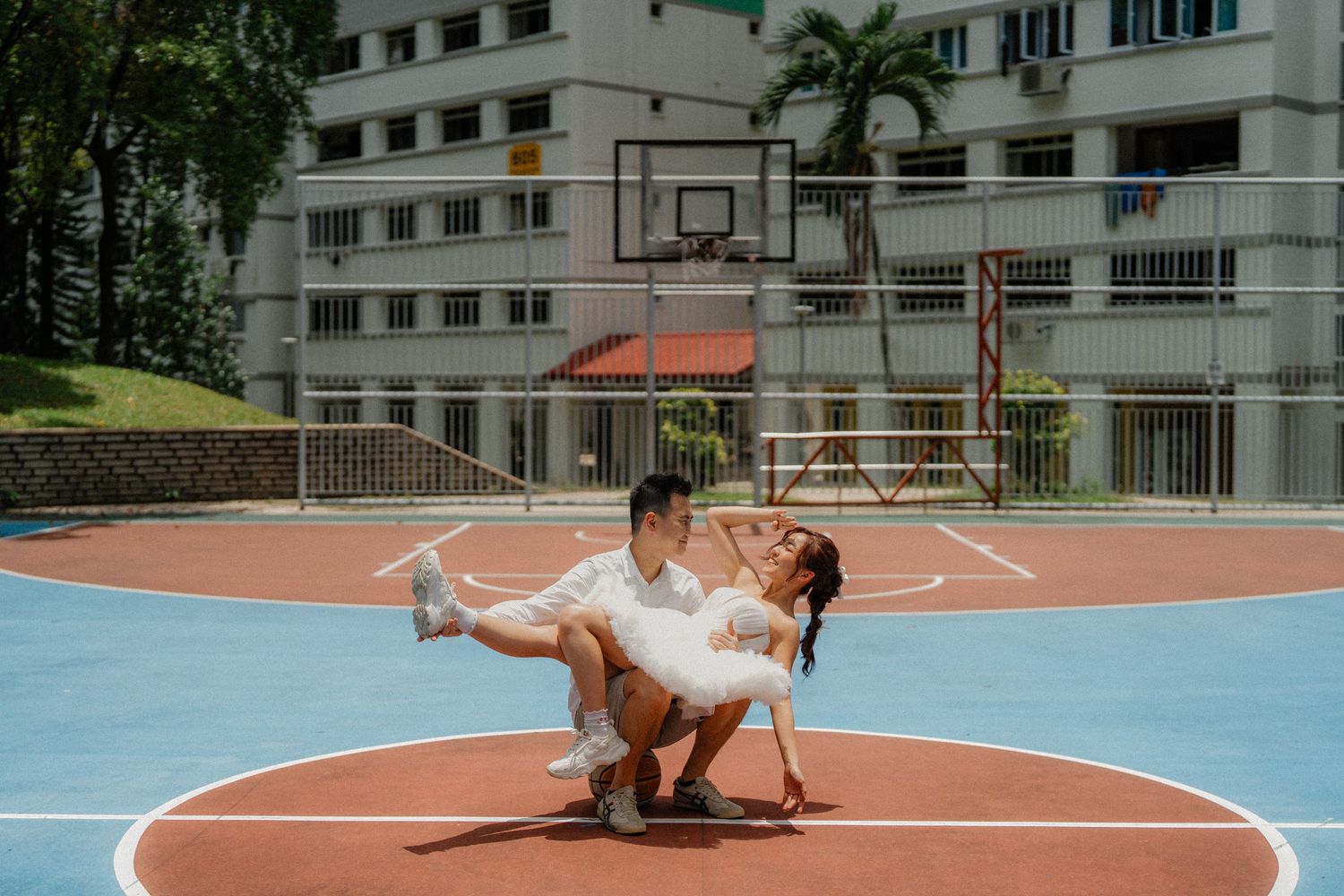 A couple in wedding attire performs a romantic dance pose on an outdoor basketball court near an apartment building.