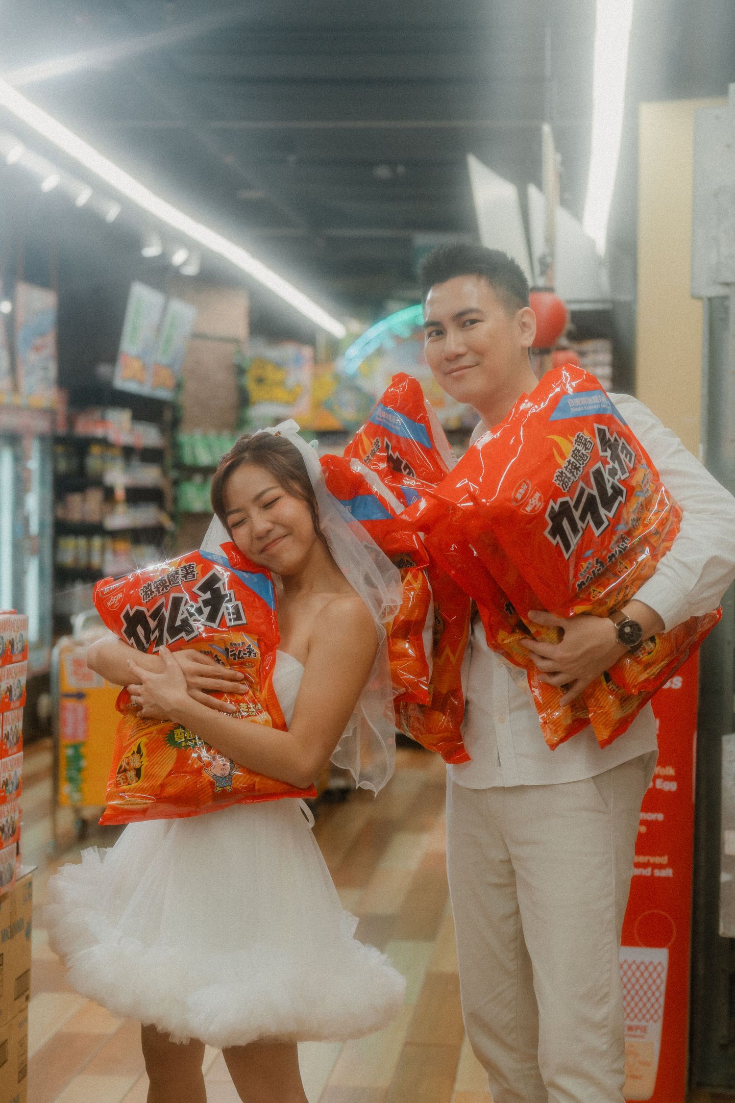 A couple in wedding attire poses playfully with snack bags in a grocery store aisle.