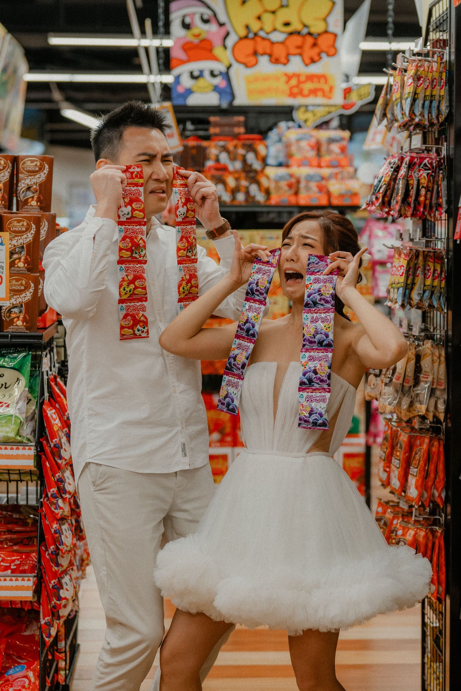 Playful couple holding snack packages in front of colorful store displays.