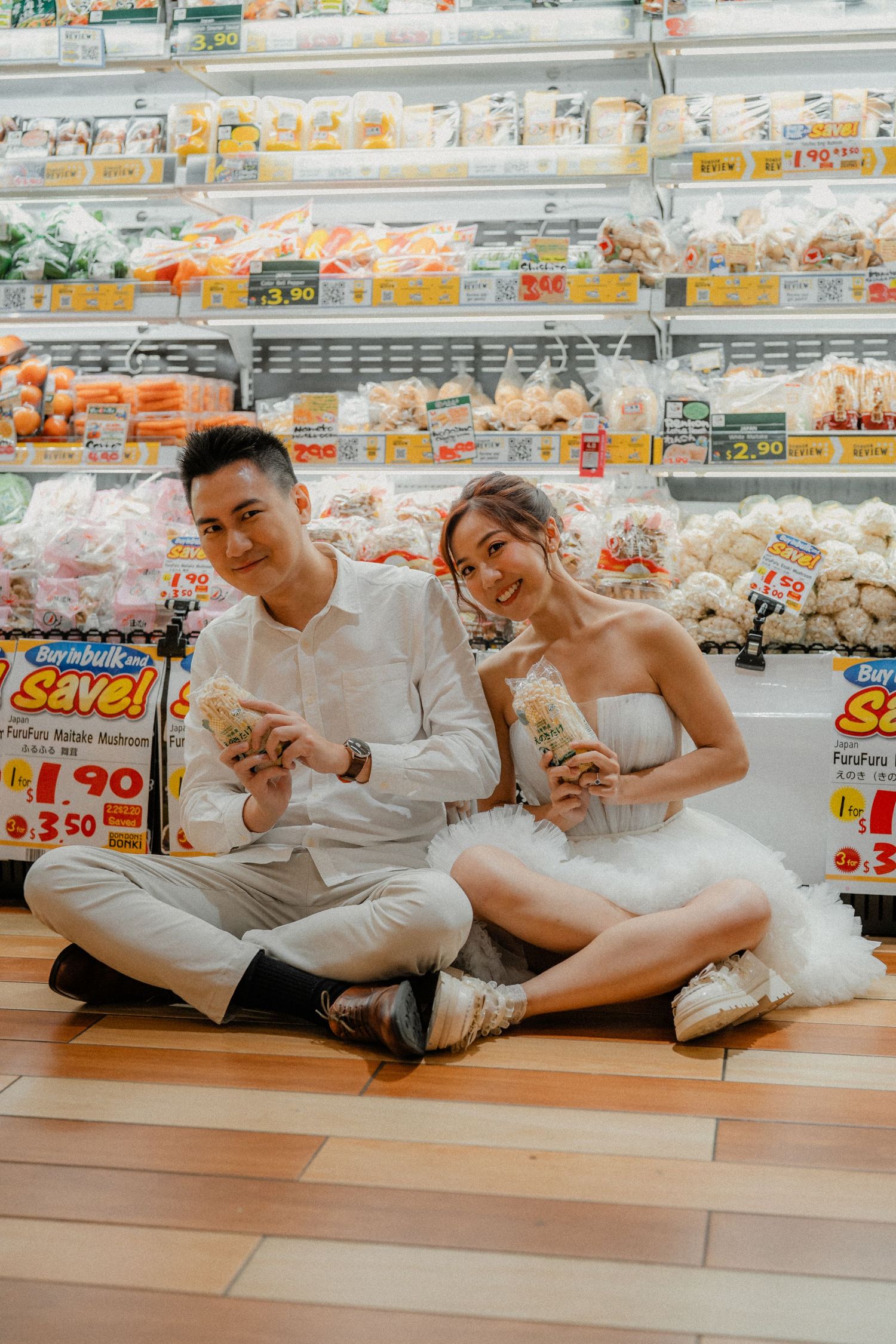 Couple sits on grocery store floor sharing snacks while dressed in elegant white attire.