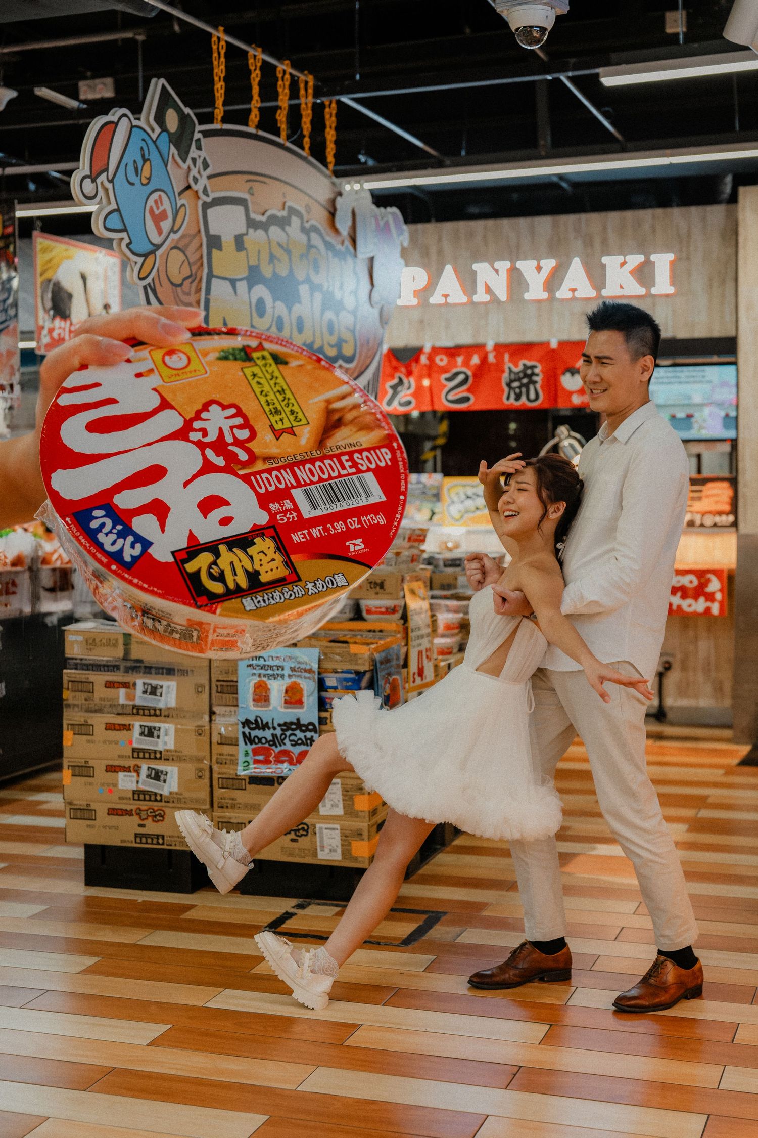 Playful couple pose with Japanese snack banners in a grocery store, creating whimsical dance-like movements.