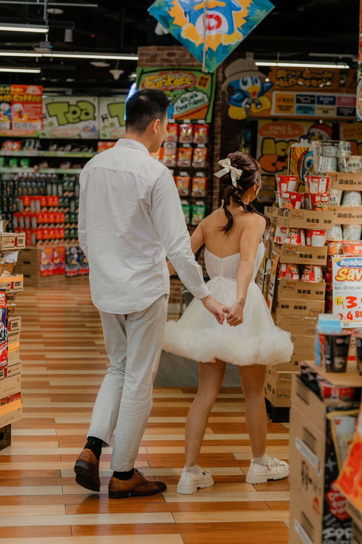 Couple in white attire walks through grocery store snack aisle holding hands.