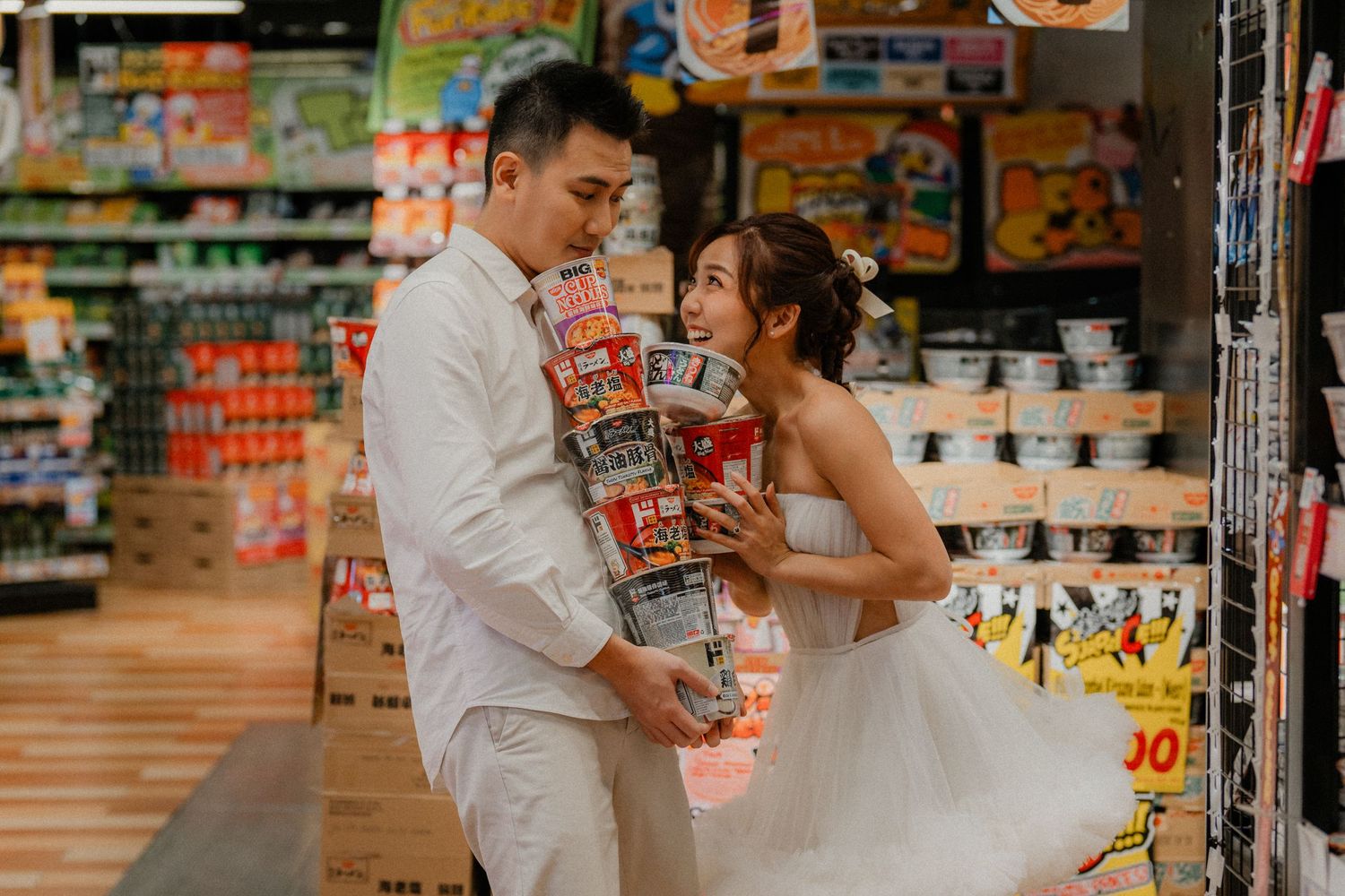 Wedding couple playfully posing with stacked snacks in grocery store aisle.