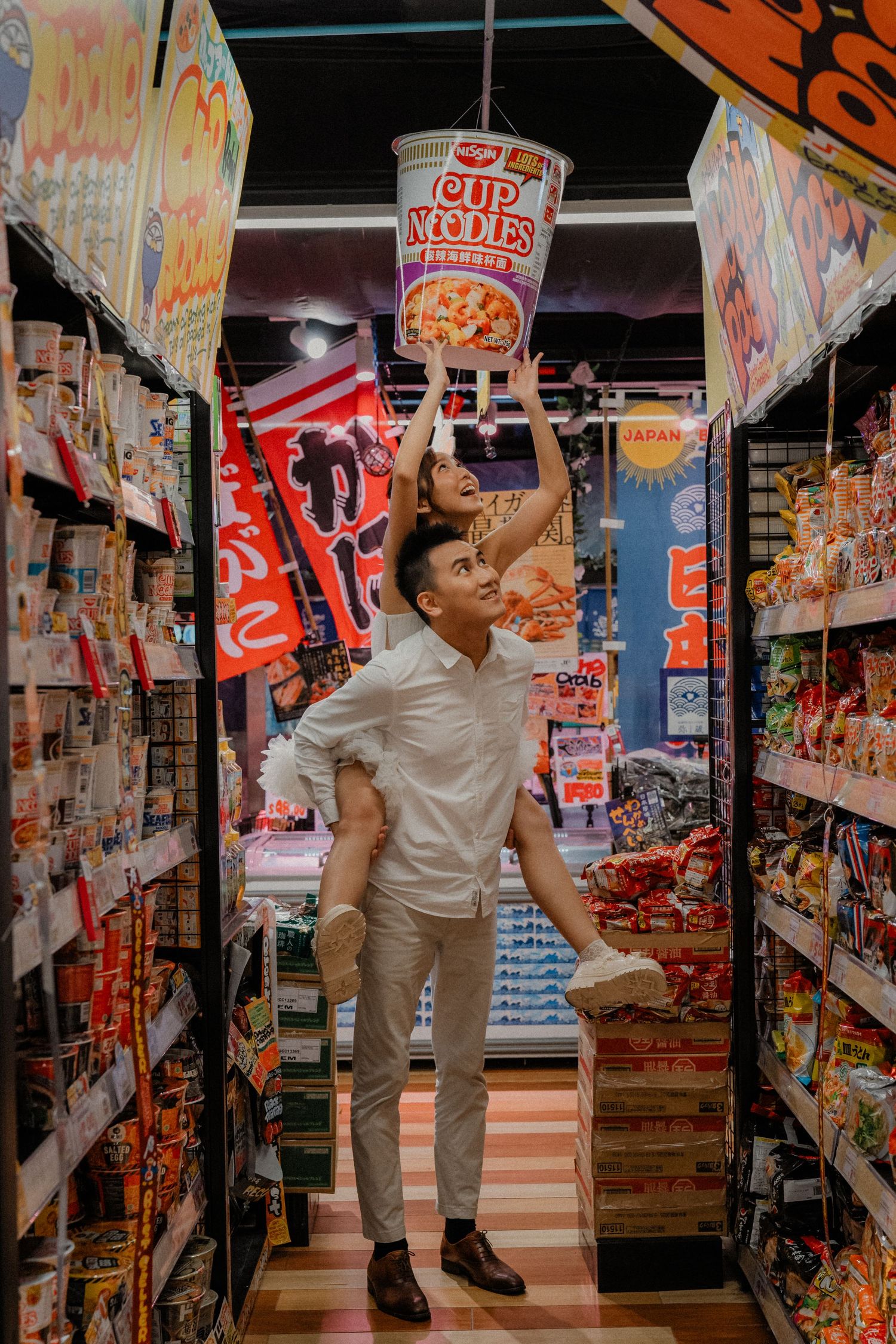 A couple poses playfully in a store aisle filled with colorful snacks and food products.
