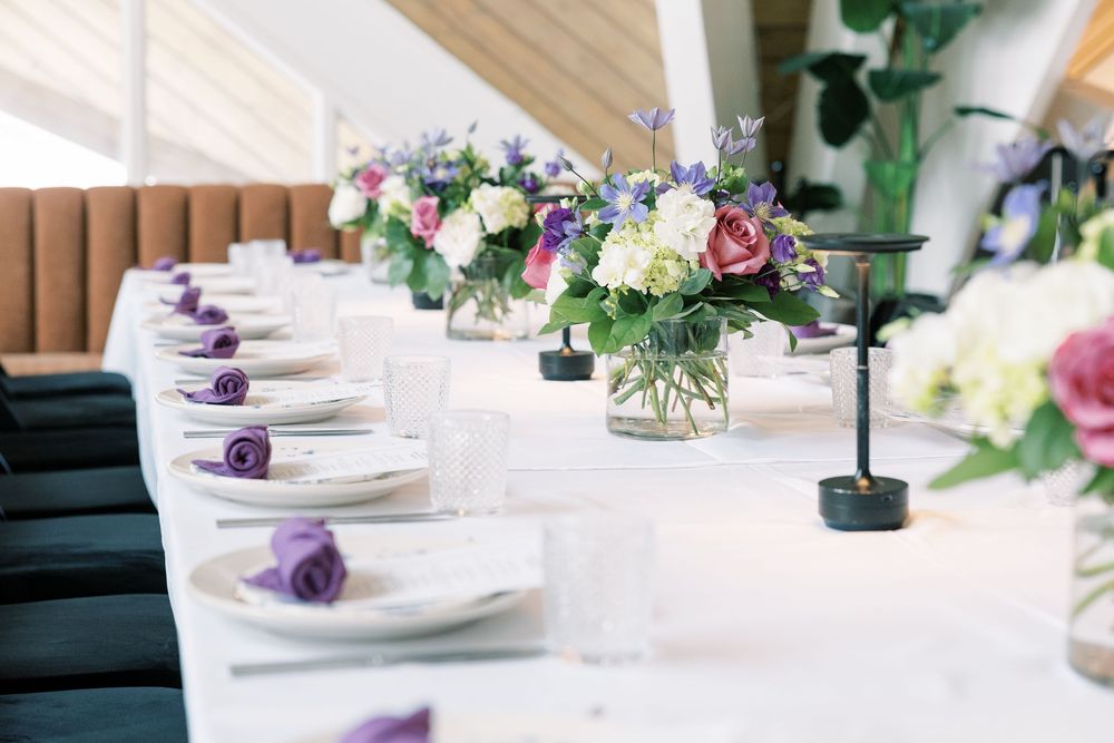 An elegant table setting with white plates, purple accents, and fresh floral centerpieces in glass vases on a white tablecloth.
