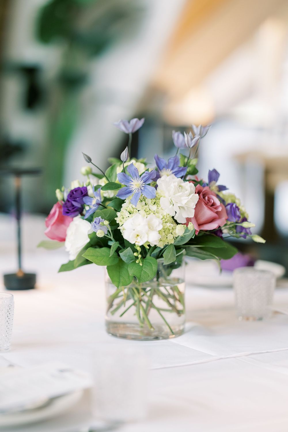 A delicate floral centerpiece with pink roses, white hydrangeas and purple blooms in a glass vase on a white tablecloth.