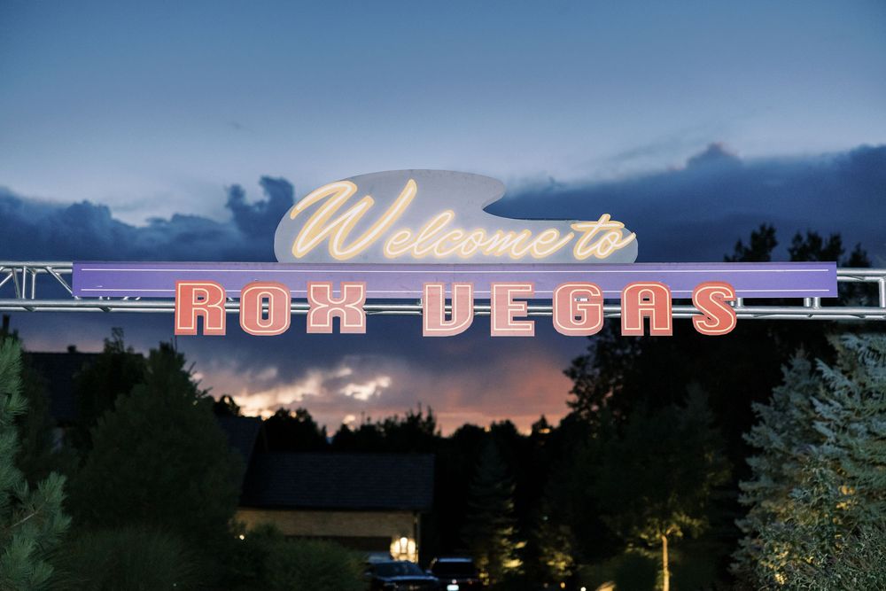 Glowing pink neon sign spelling 'Mon Vegas' illuminated against a dusky blue sky with trees silhouetted below.