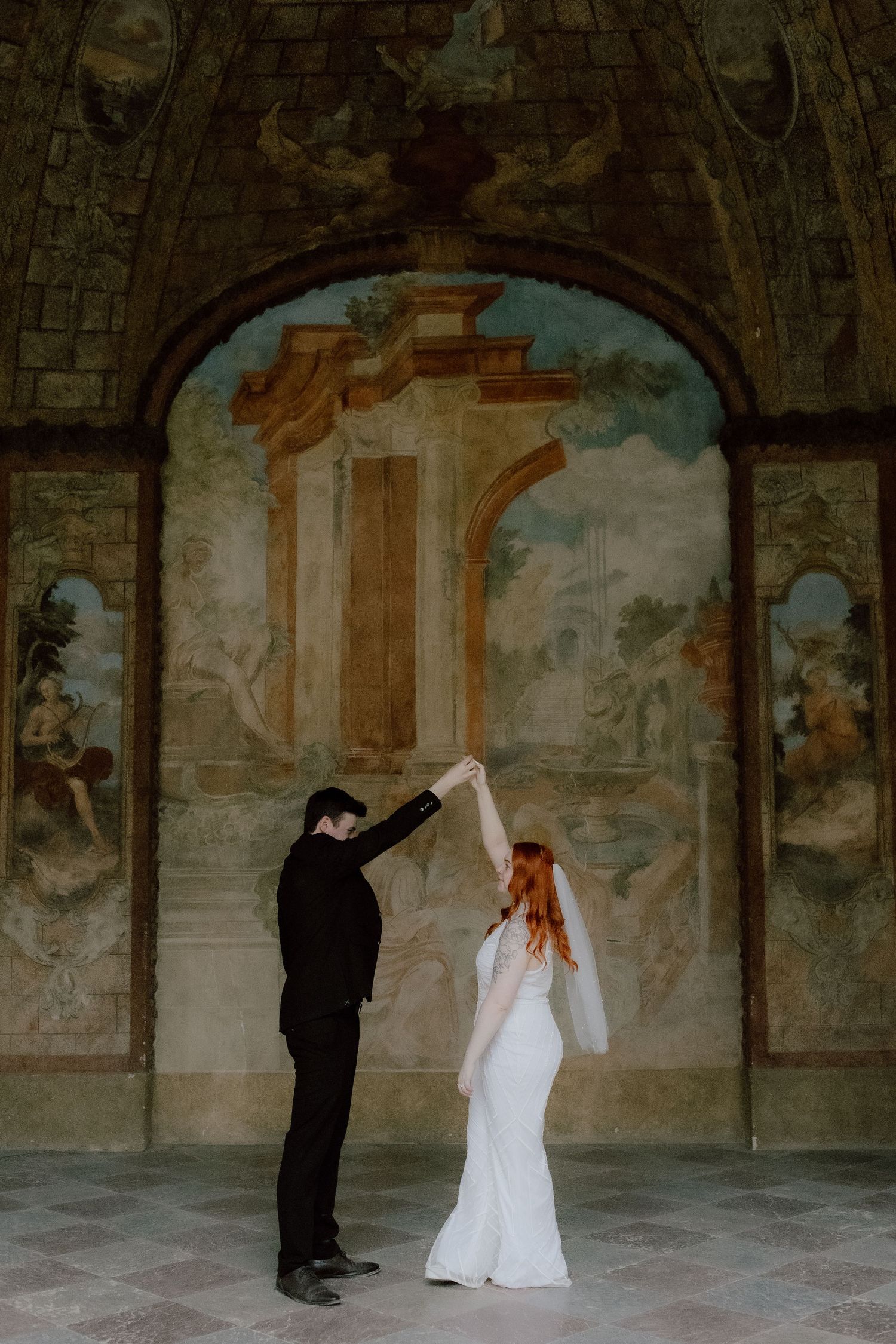 A couple in wedding attire dancing together under an ornate archway with classical fresco paintings and architecture.