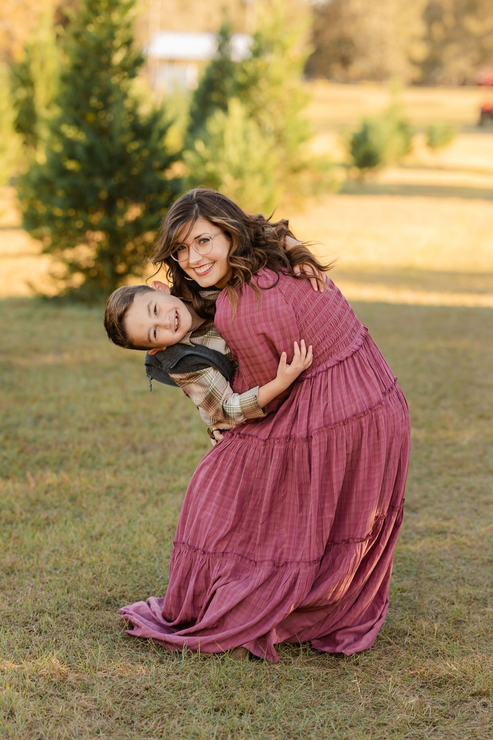 Two people share a joyful embrace outdoors while one wears a flowing mauve dress in a golden-lit field with evergreens.