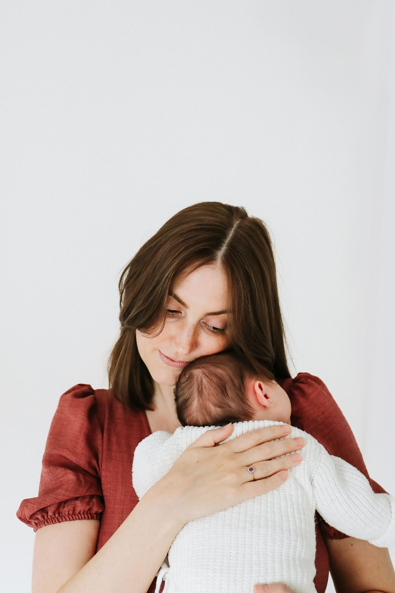 A tender moment between mother and newborn baby in white clothing against neutral background, during newborn shoot in Nottingham