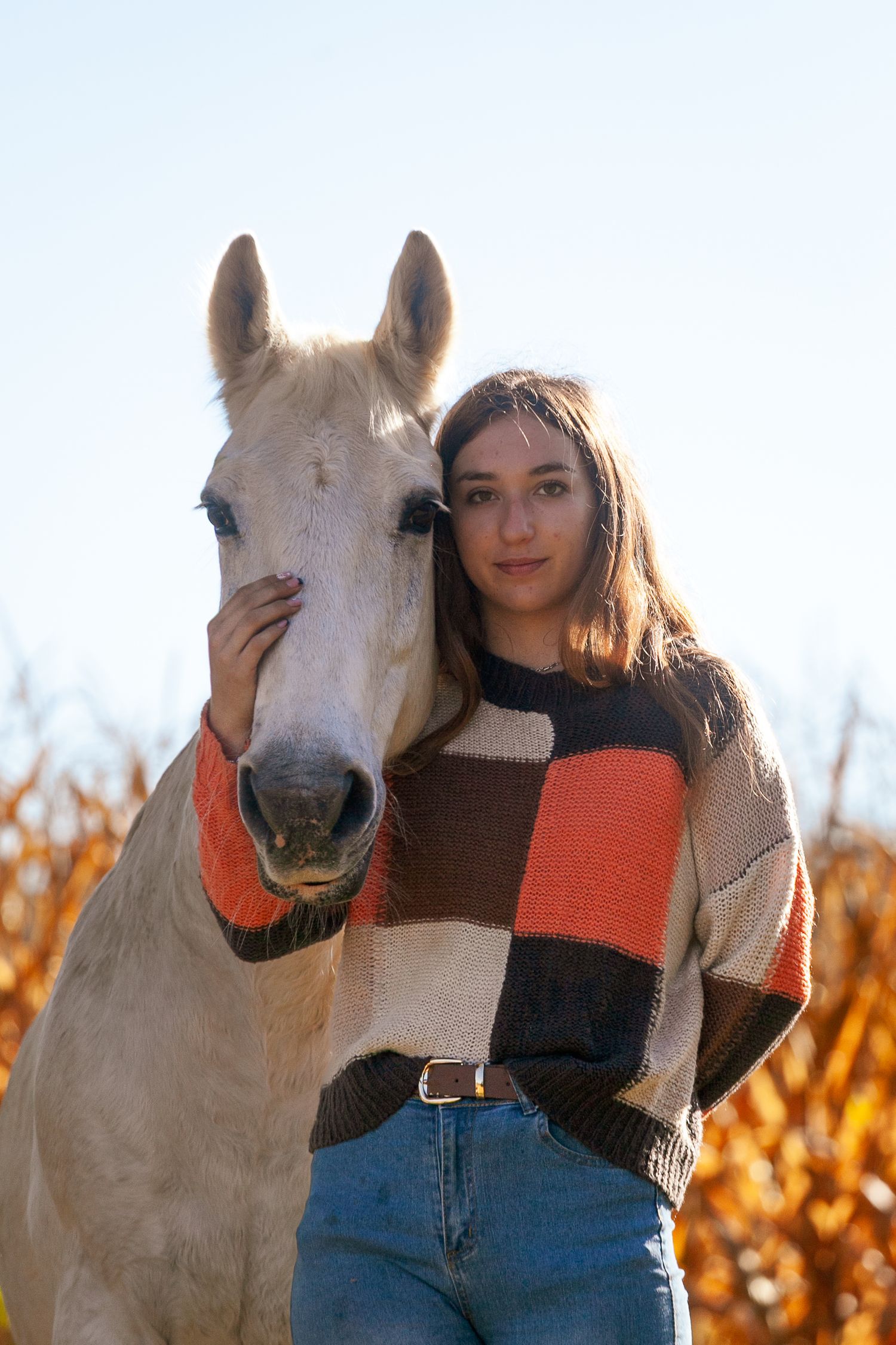 A white horse poses with someone wearing a brown sweater against an autumn backdrop.