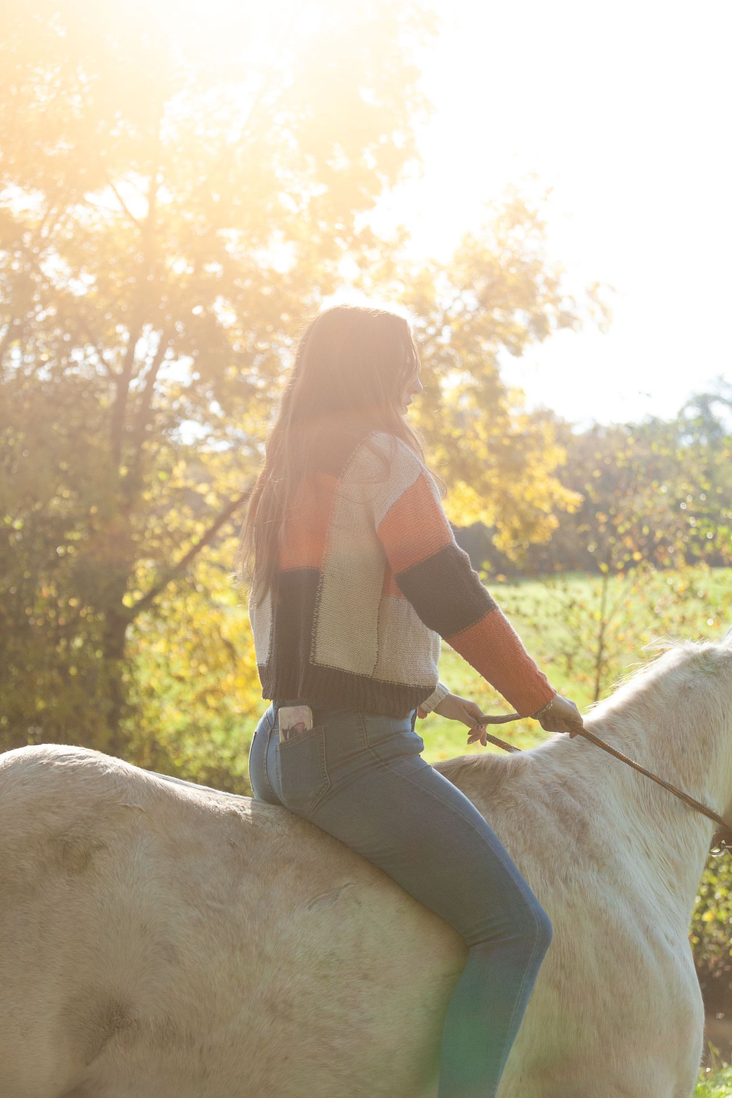 Person riding bareback on a white horse during sunset with golden light filtering through trees.