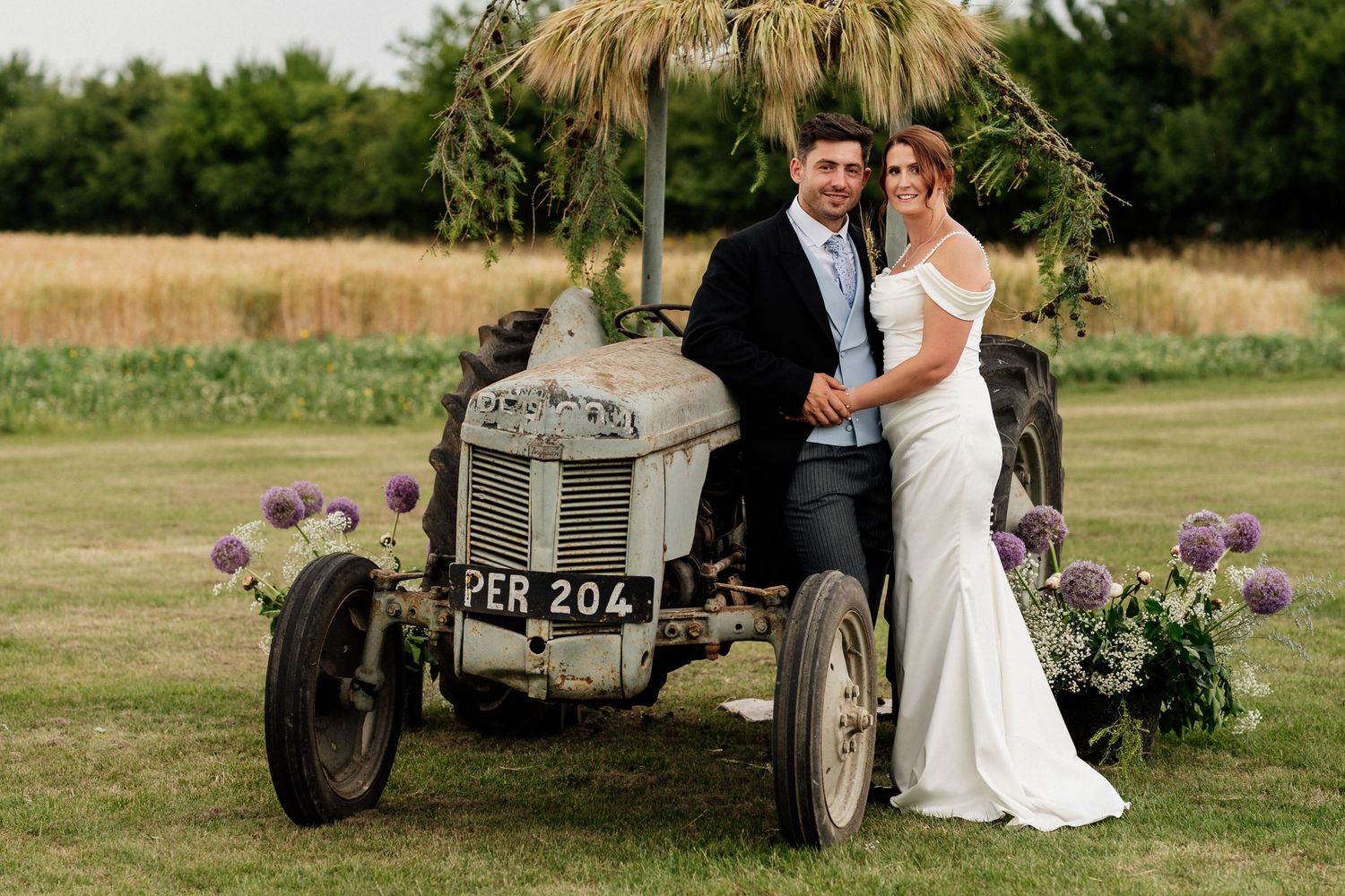 Romantic wedding couple sitting beside vintage gray tractor in rustic countryside setting at sunset.