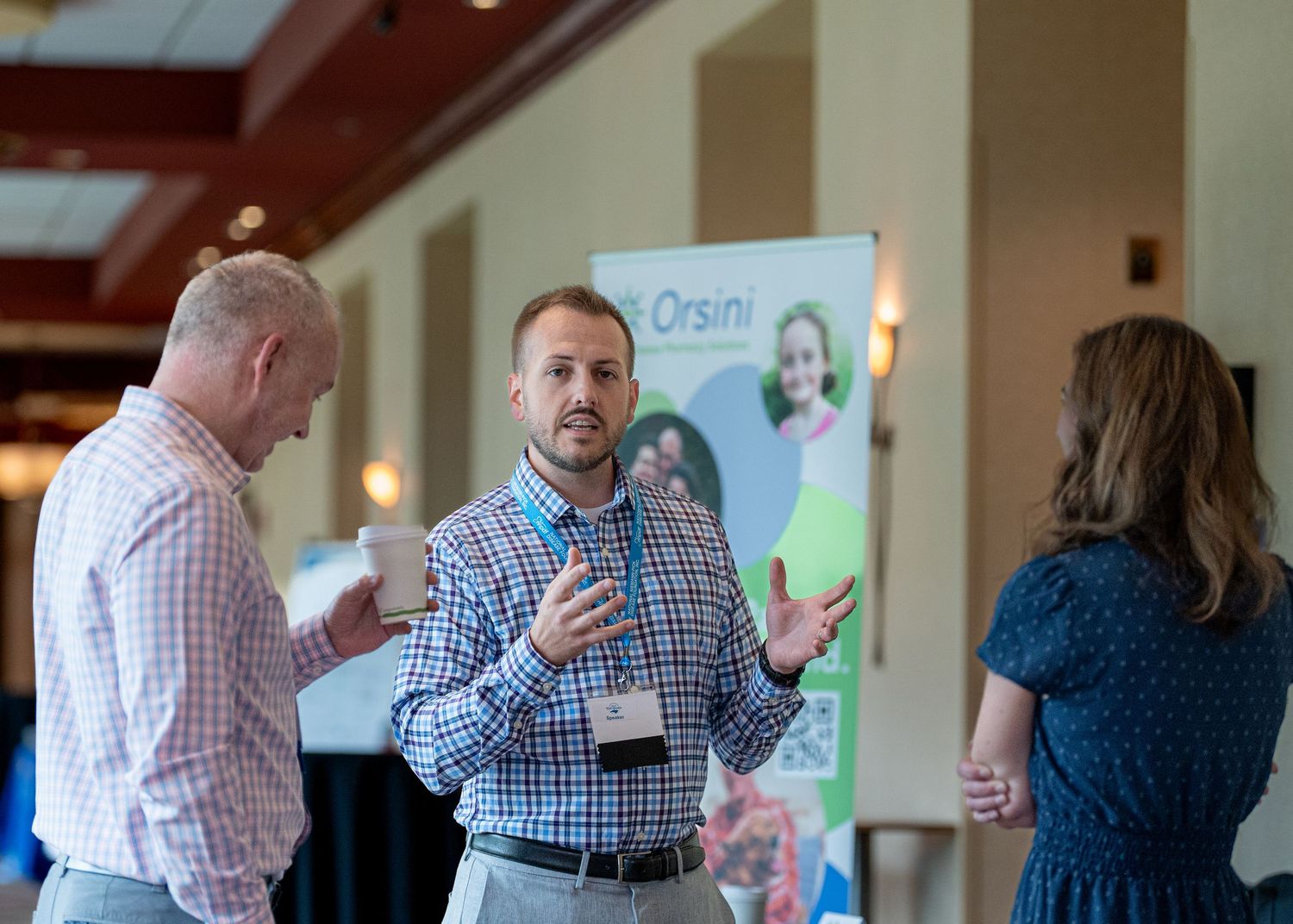 Three people engaged in animated conversation at a professional conference with banners visible in the background.