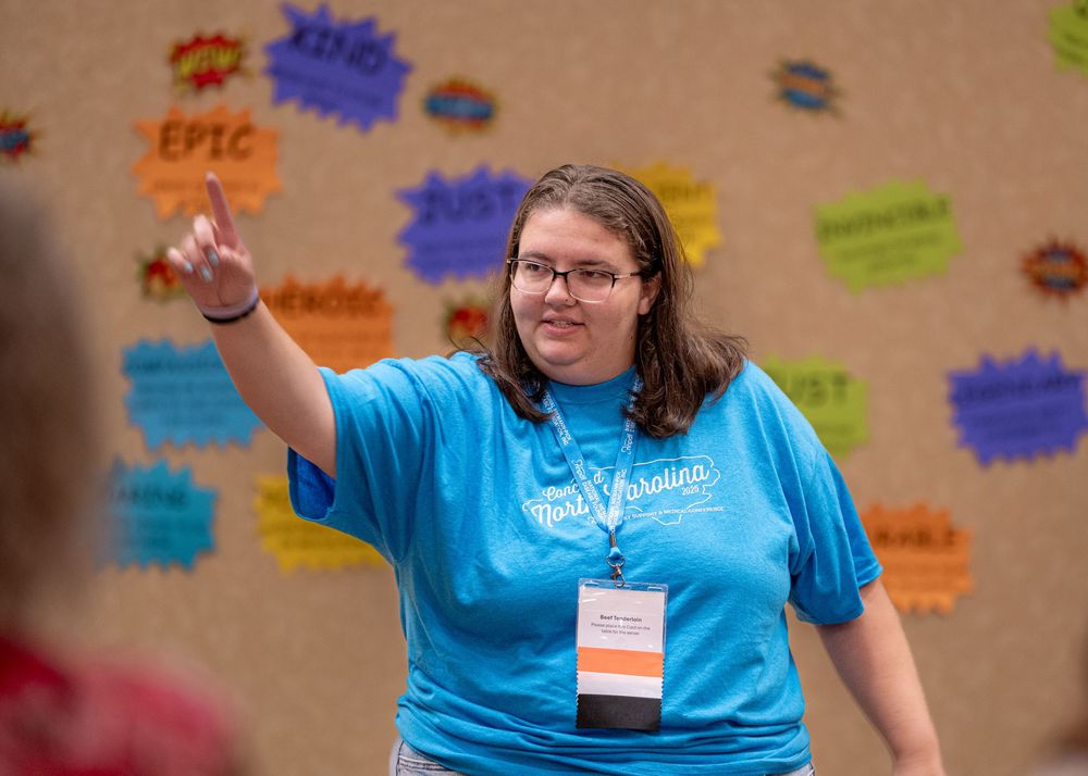 Event participant in bright blue shirt gesturing while speaking in front of a colorful bulletin board.