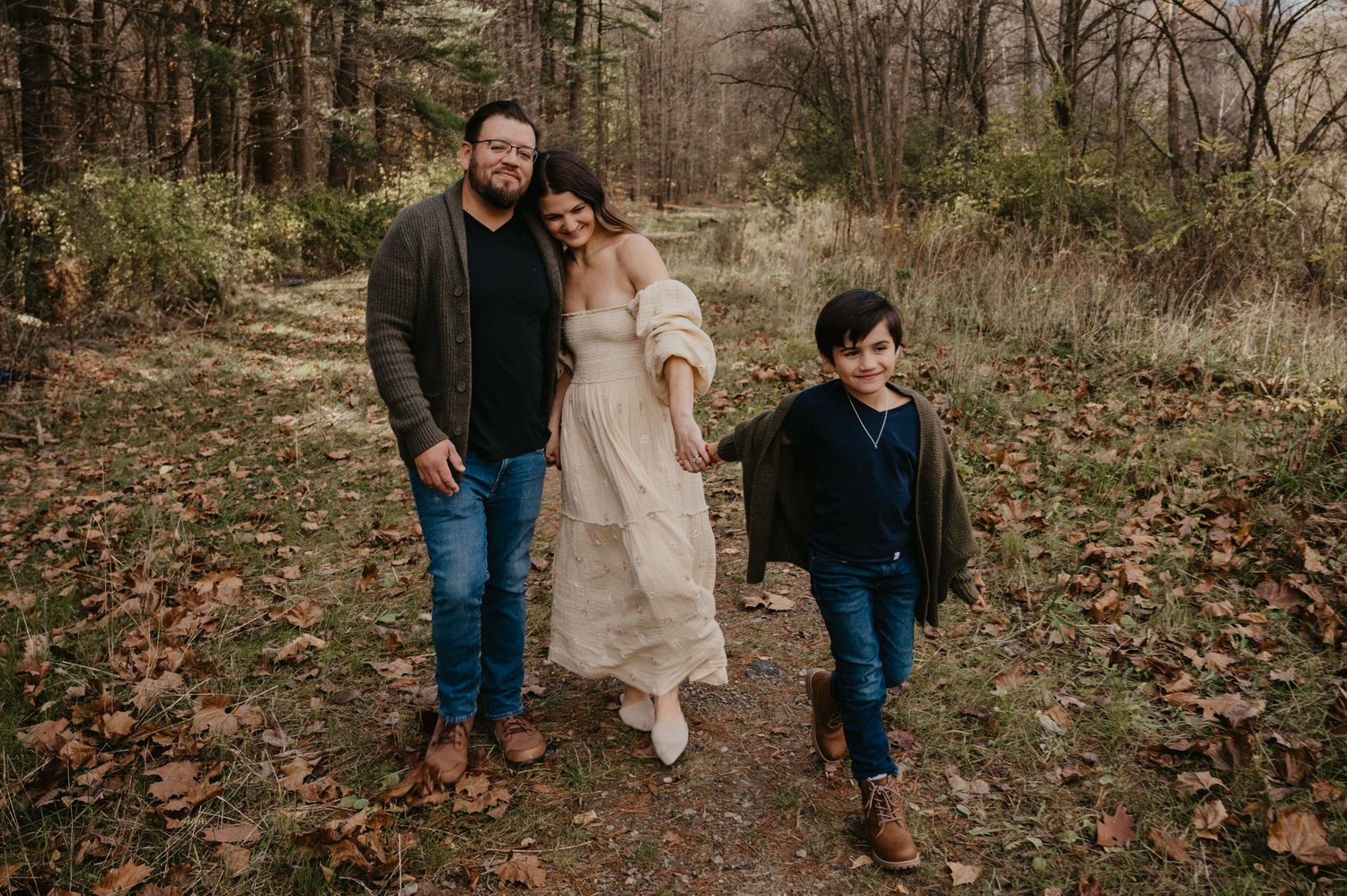 Family of three walking together on a forest path in autumn, parents holding hands with young son among fallen leaves.