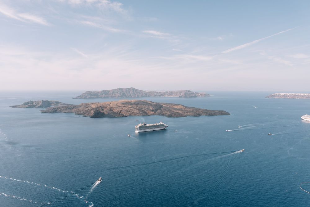 Large cruise ship anchored near volcanic islands in blue Mediterranean waters off Santorini, Greece, viewed from above.