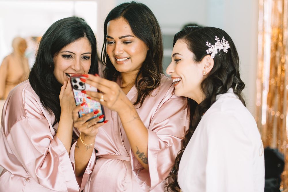 Three bridesmaids in pink silk robes look at a phone together while getting ready for a wedding celebration.
