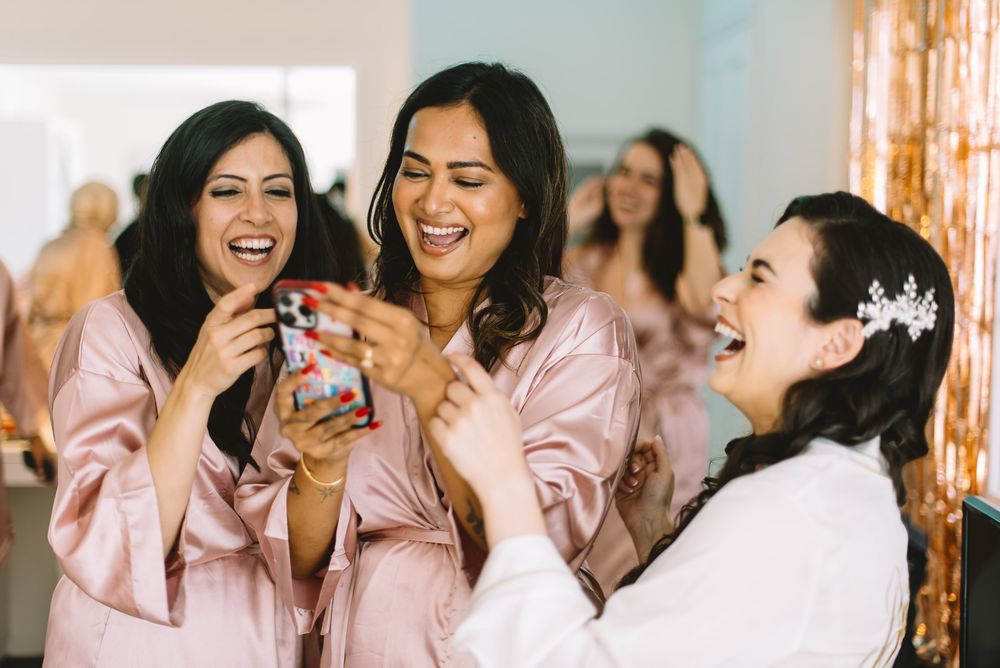 Bridesmaids in pink silk robes laugh together while getting ready before a wedding ceremony.