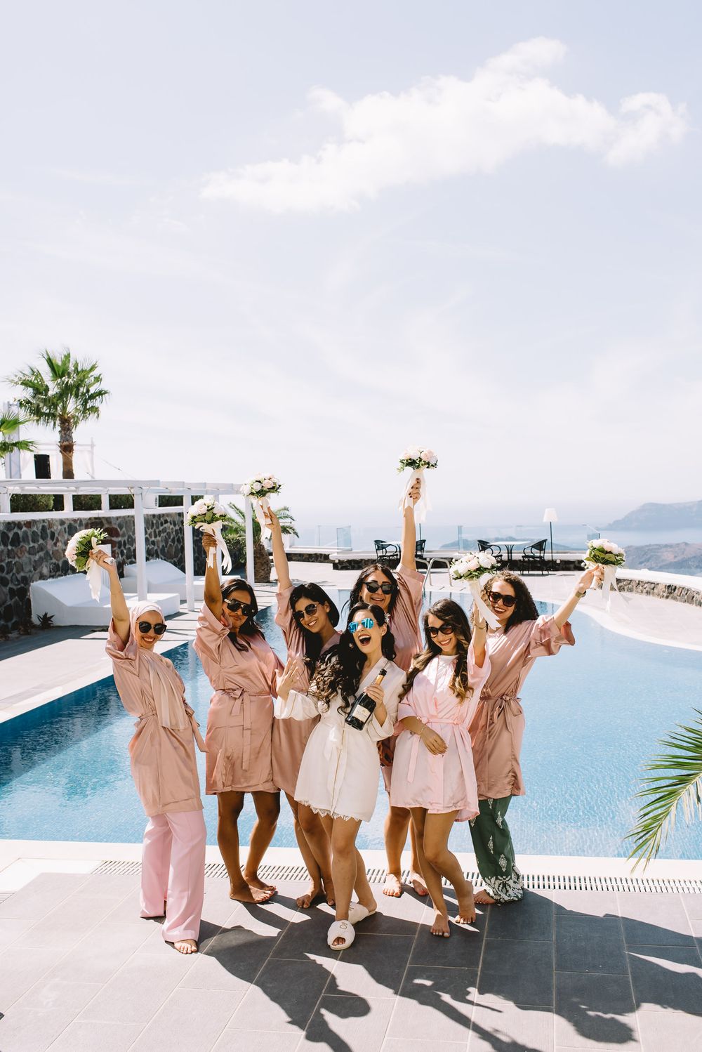 Bridal party in matching pink robes celebrates by poolside with bouquets at scenic Mediterranean destination wedding venue.