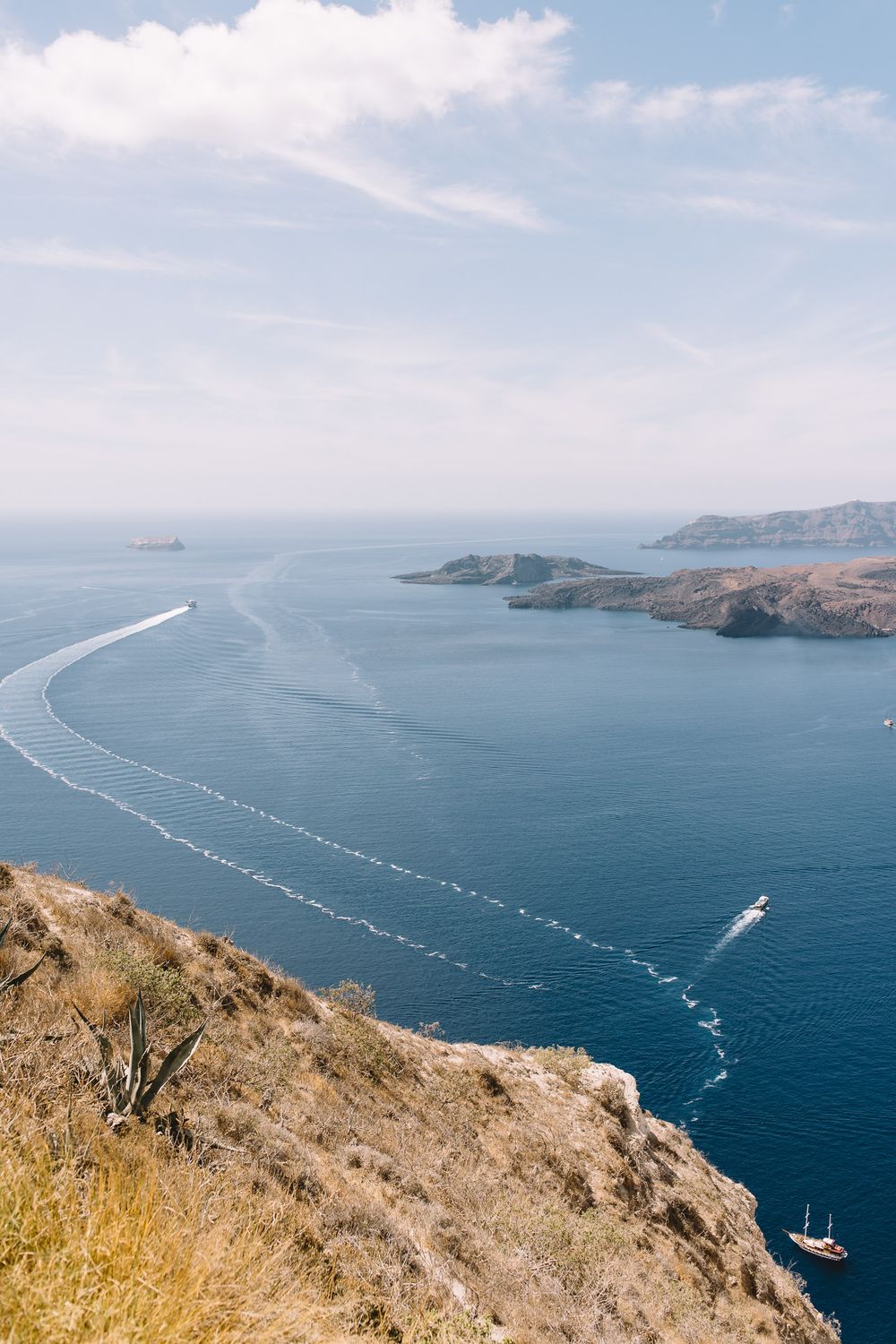 Aerial view of boats creating wake patterns in blue Mediterranean waters along rocky coastal cliffs on a sunny day.