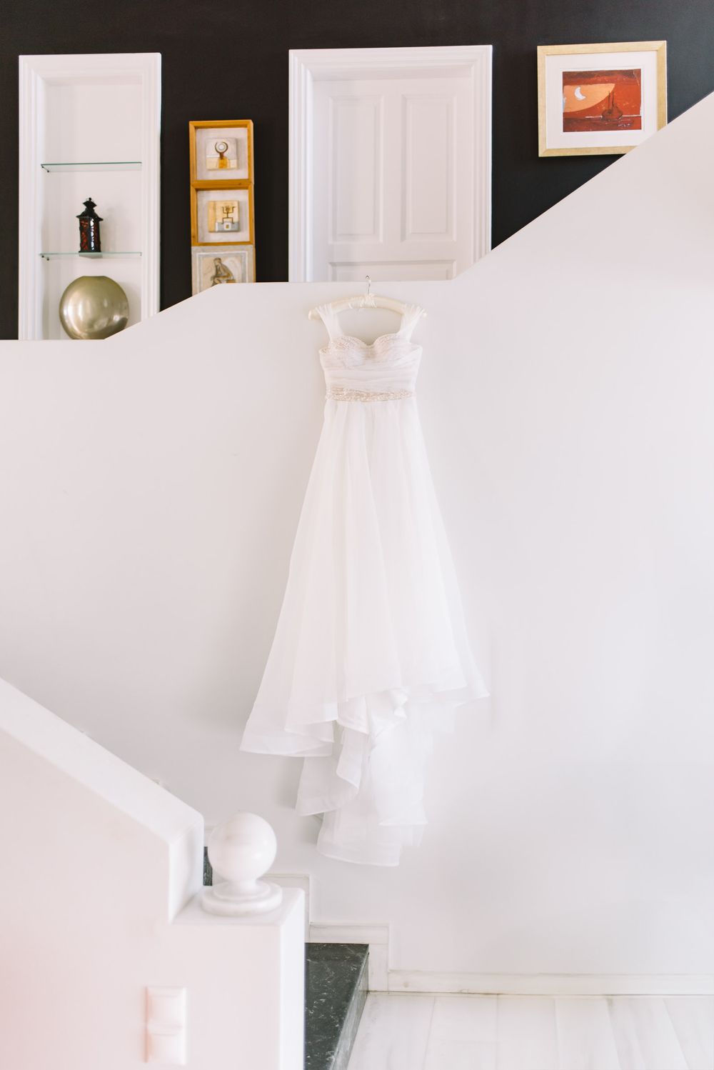 A white wedding dress hangs elegantly on display against a bright wall with framed artwork and a decorative gold sphere.