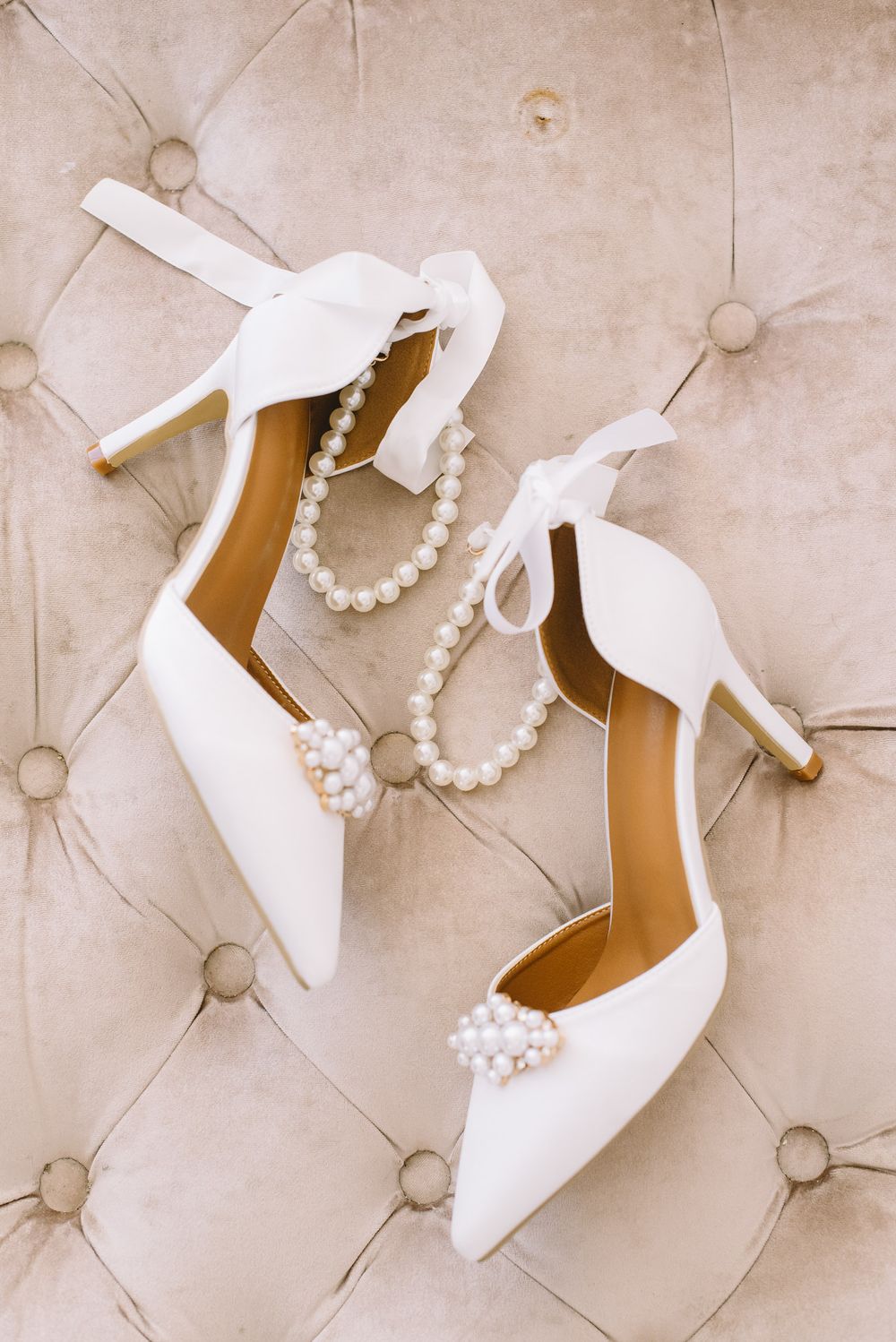 Elegant white bridal heels with pearl straps displayed on beige tufted upholstery.