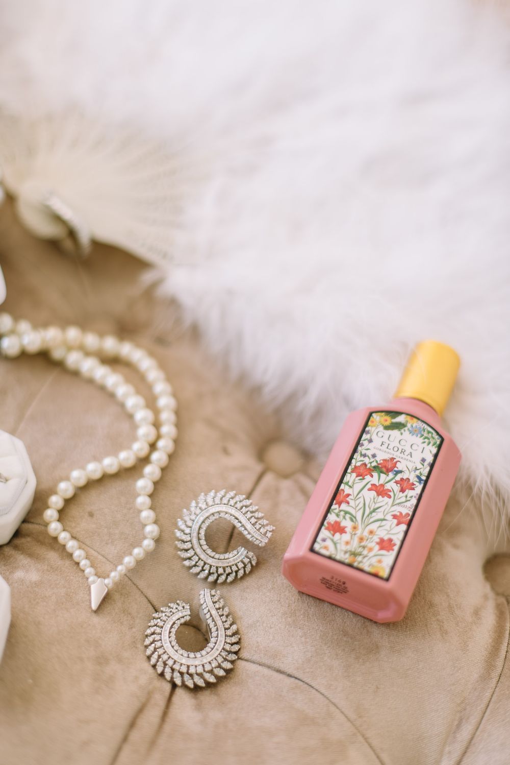 Styling essentials including pearl necklace, silver brooch and pink perfume bottle arranged on beige fabric.