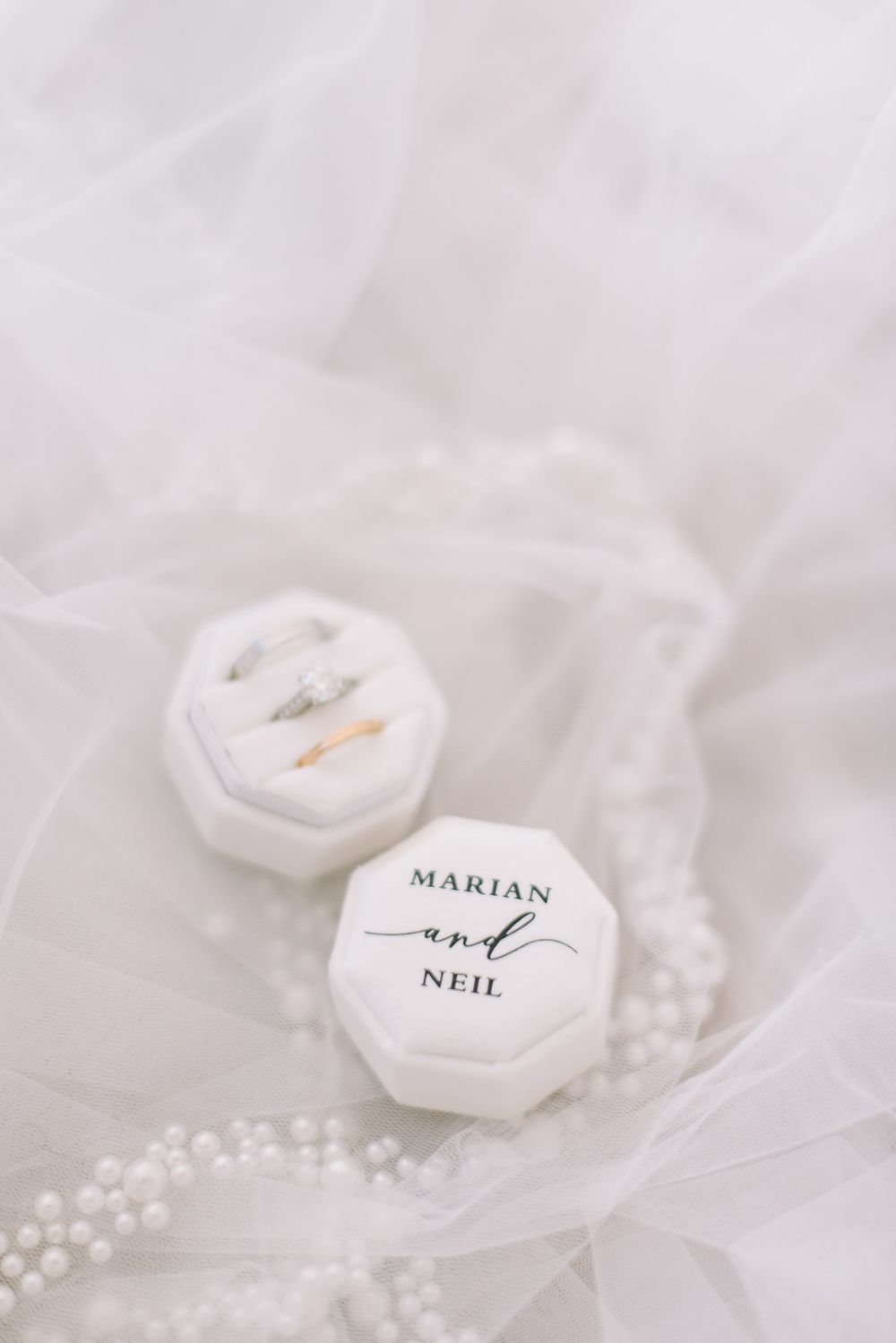 White hexagonal ring boxes with couple's names and wedding rings resting on delicate tulle fabric with pearl details.