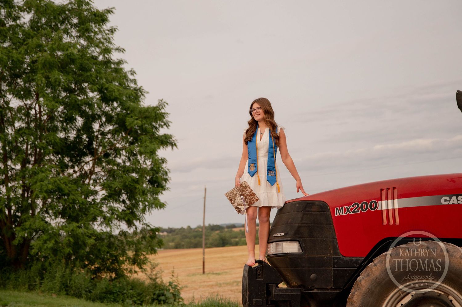 Standing on a red Case tractor wearing a white summer dress and blue scarf in a rural farm setting.