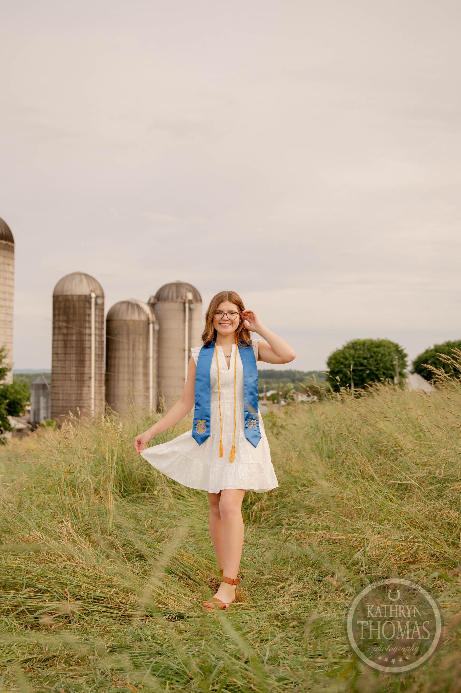 A white dress and denim jacket outfit is showcased against a rural farm backdrop with tall silos and grassy fields.