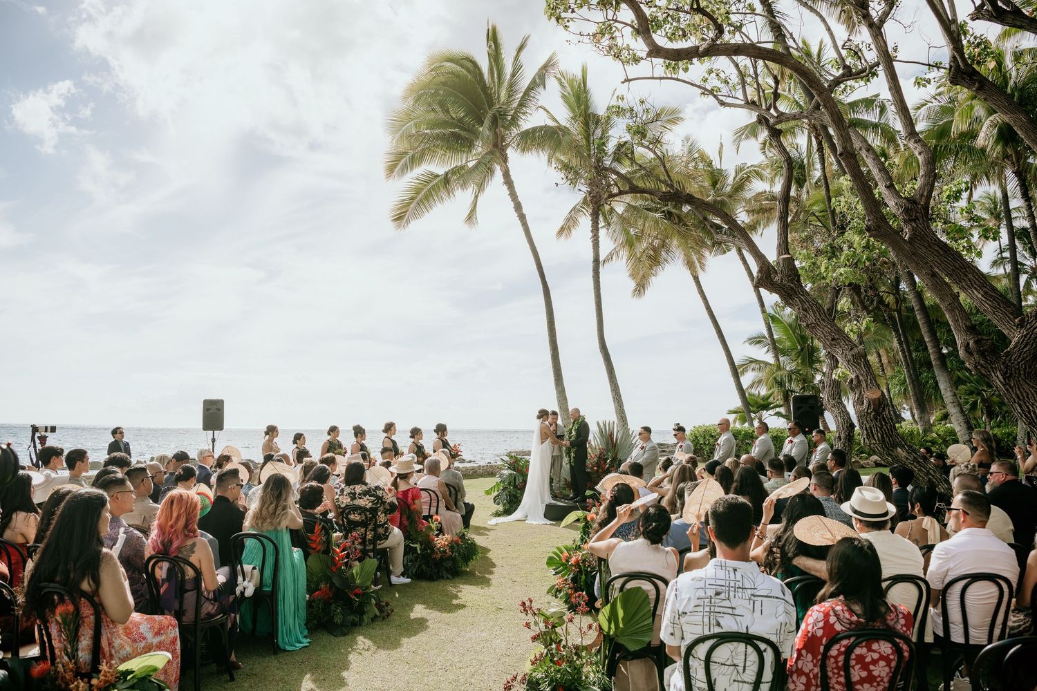 A beachside wedding ceremony under swaying palm trees with guests seated in white chairs facing the ocean.