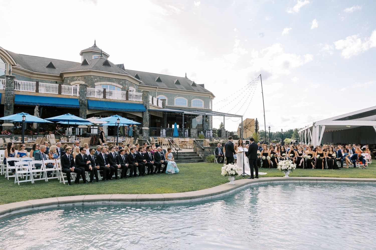 A panoramic view of an outdoor graduation ceremony with rows of attendees seated under white tents near a school building.