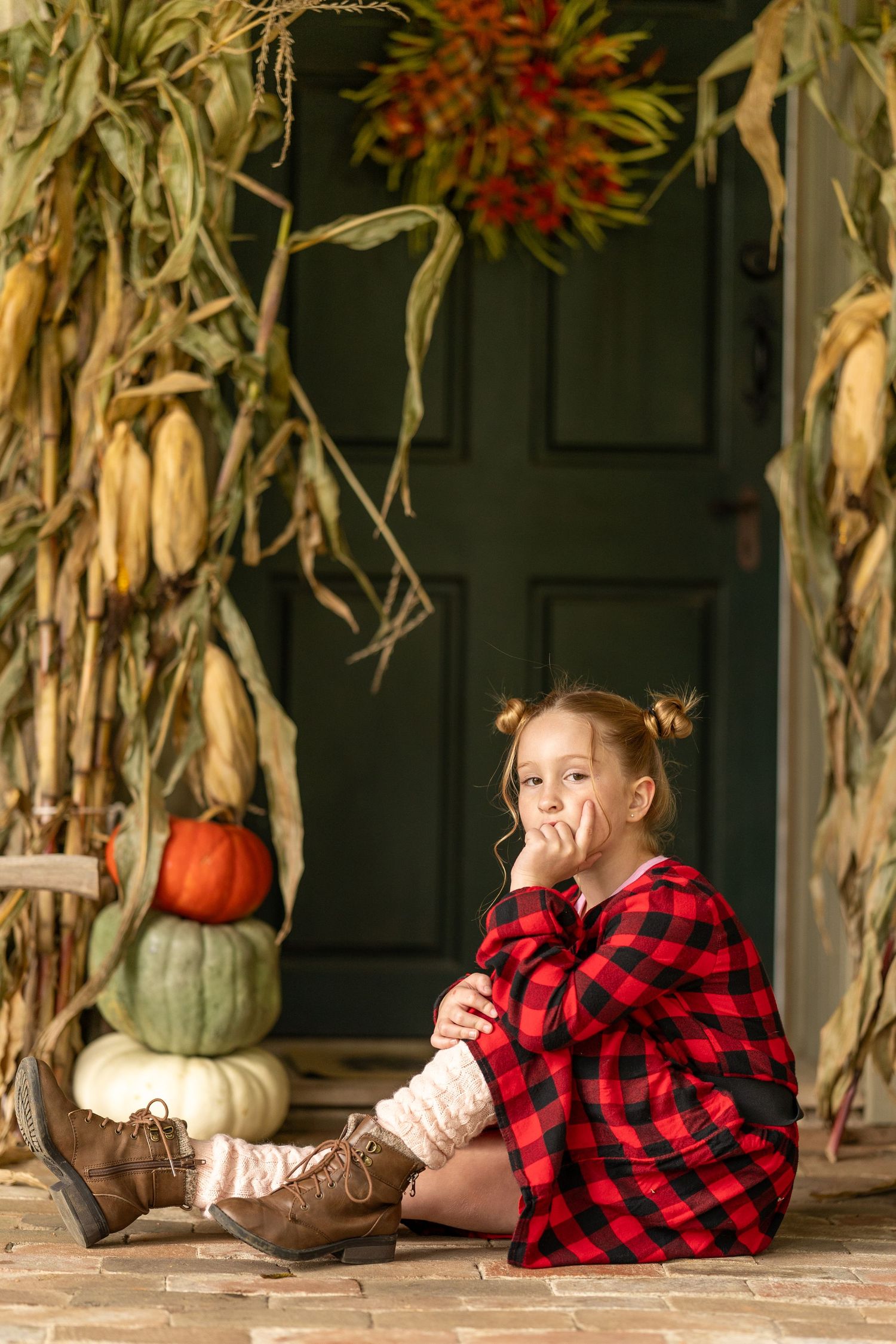 Child in buffalo plaid sitting on porch with fall decorations including pumpkins and corn stalks. Child has hair in bun pigtails.