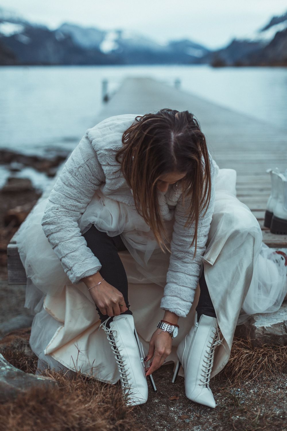Someone in a white puffer jacket and dress ties white lace-up boots by a misty mountain lake dock on a moody day.