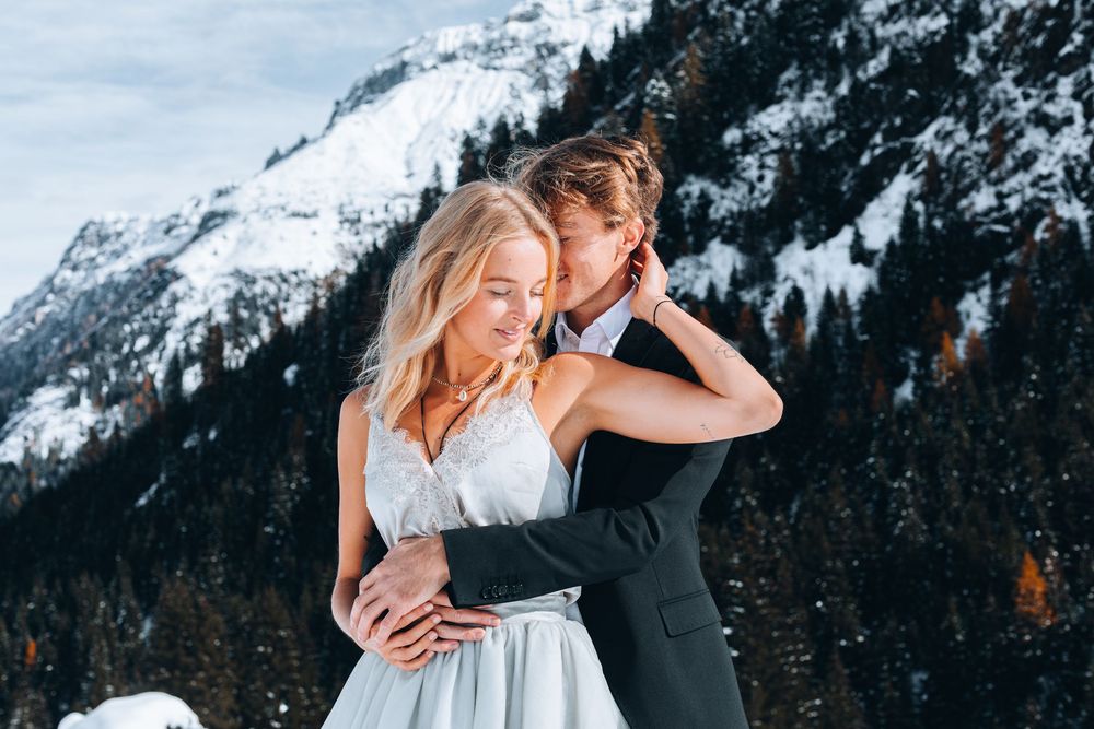 A romantic couple embraces in front of snowy mountains during a winter wedding photoshoot.