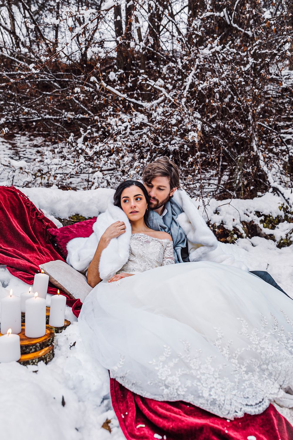 A couple lies together in fresh snow with a red fabric and white dress creating a romantic winter scene.