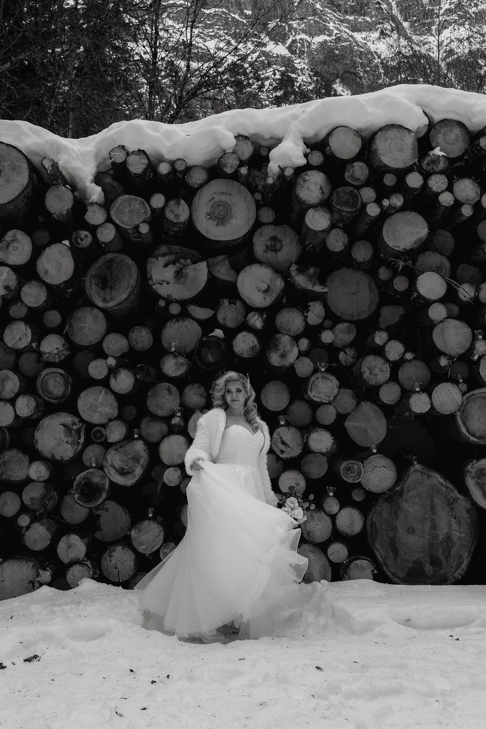 Winter wedding scene with a bride in a white dress standing against a snow-covered pile of stacked logs.