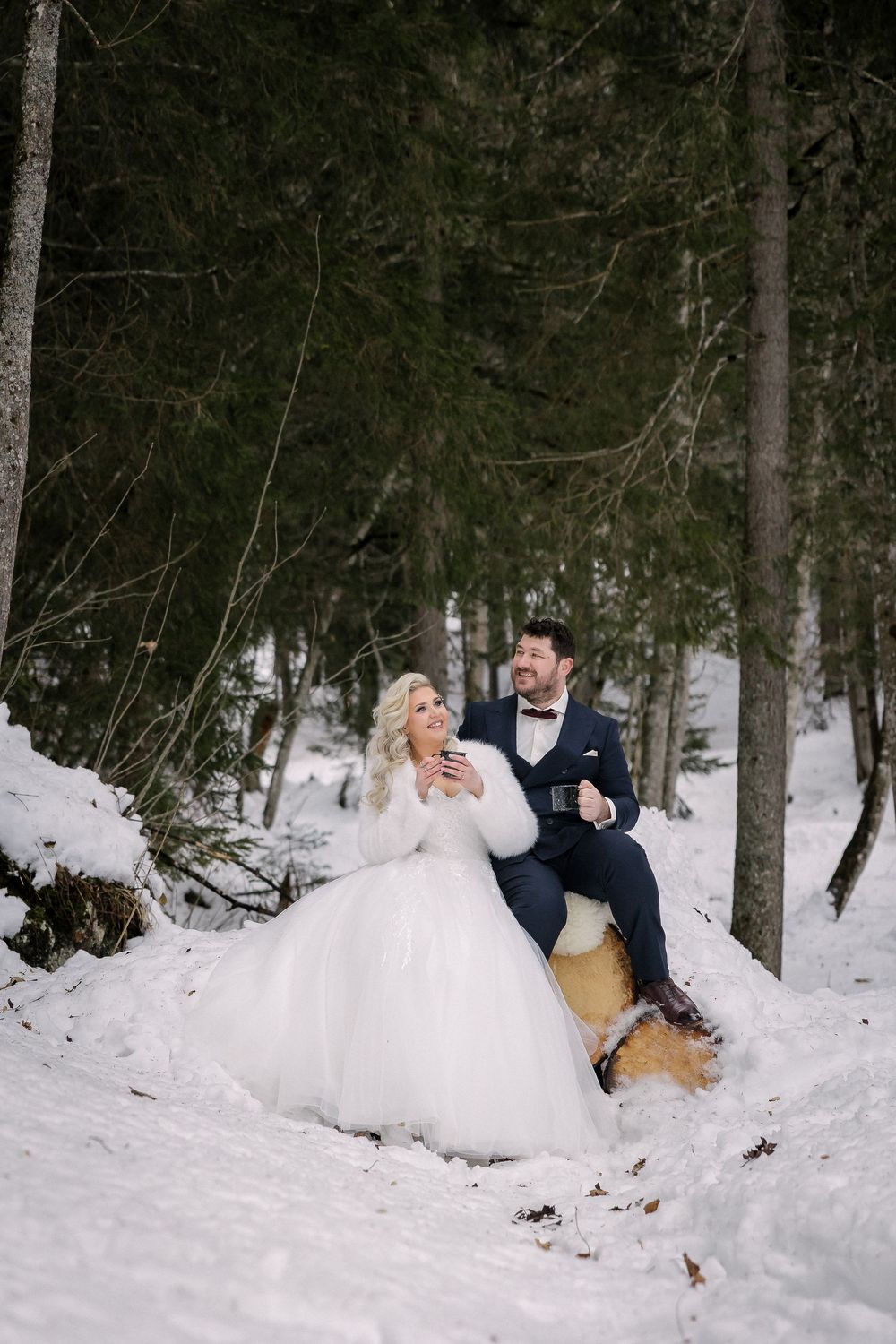 A playful winter wedding couple falls into deep snow during a photo session in a forest setting.