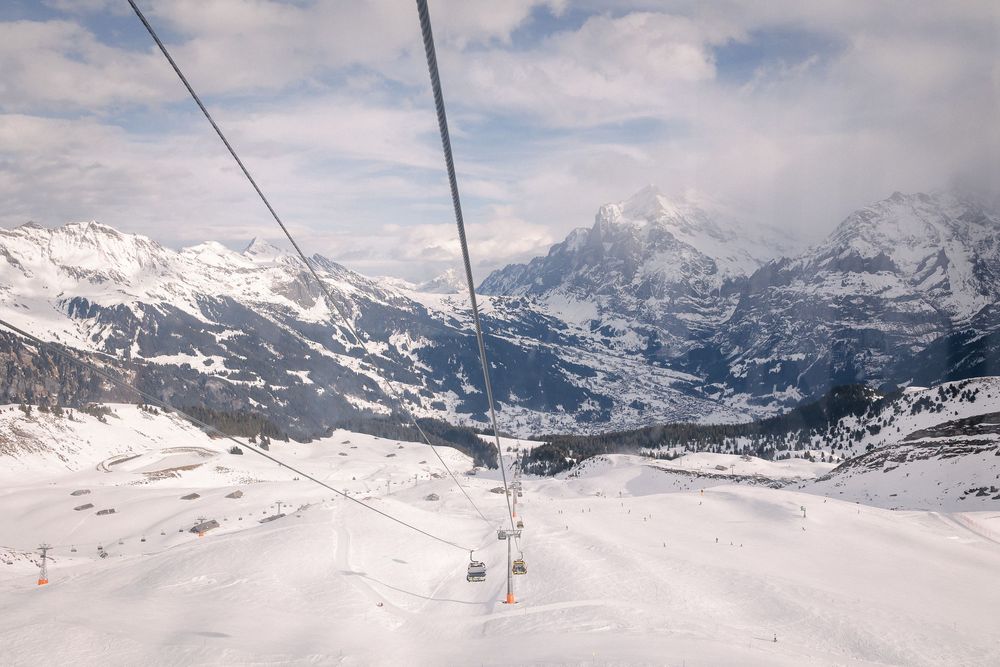 Panoramic winter landscape view of snow-covered mountain peaks and ski lift cables against a cloudy blue sky.