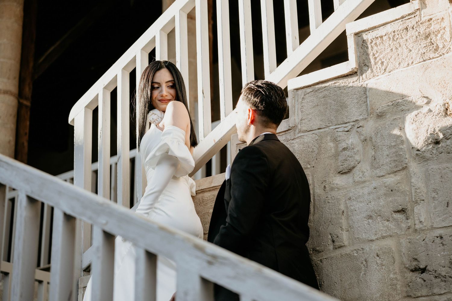 Wedding party laughs and poses on a grand white staircase in a historic stone building.