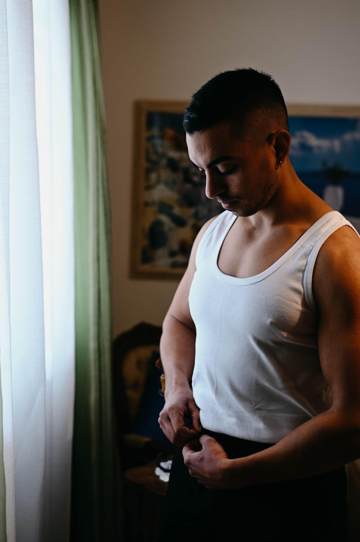 Person in white tank top standing by window in moody lighting looking down during quiet moment.