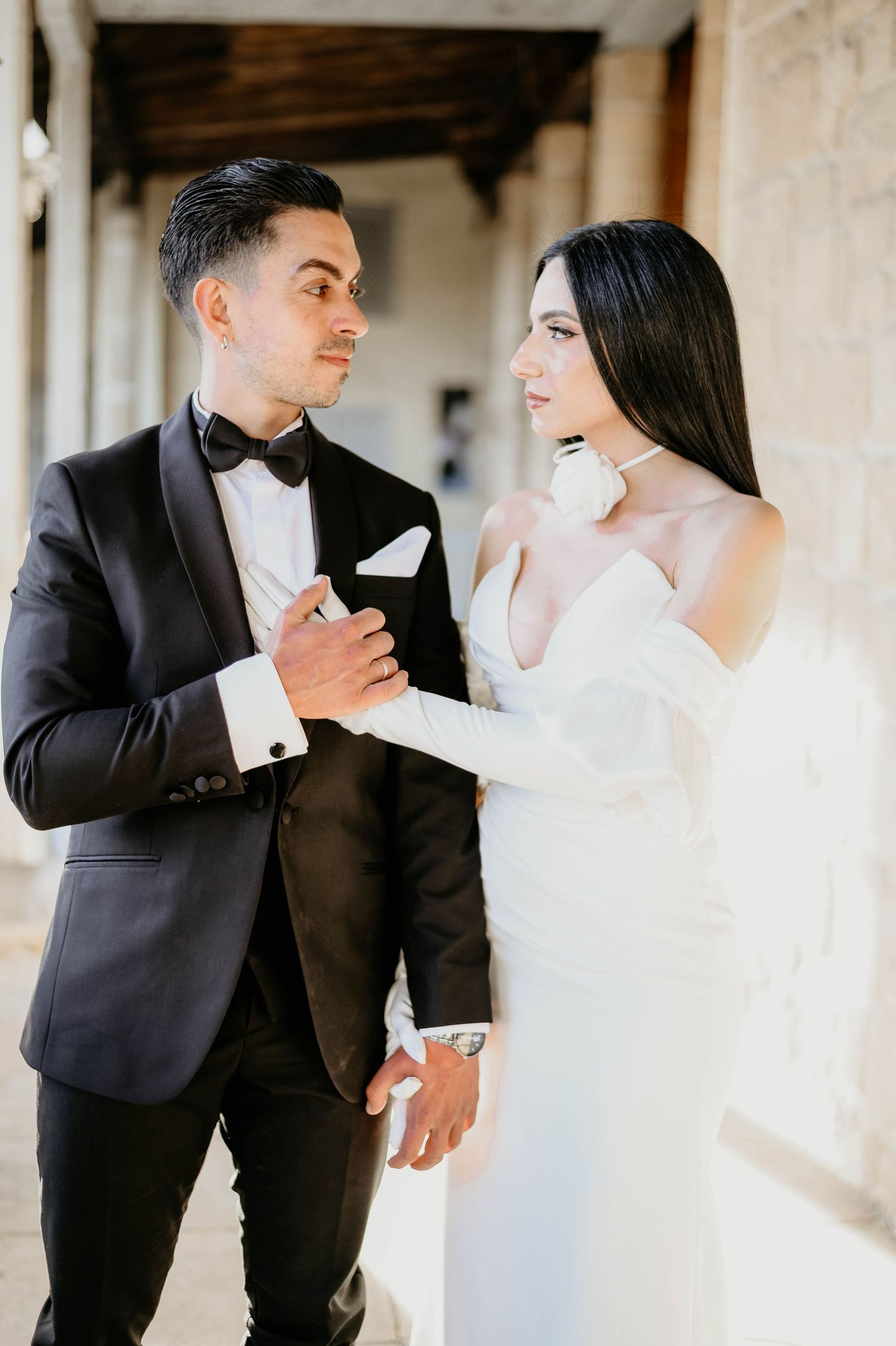 A bride in a white dress and groom in black tuxedo pose together during their elegant wedding photoshoot.
