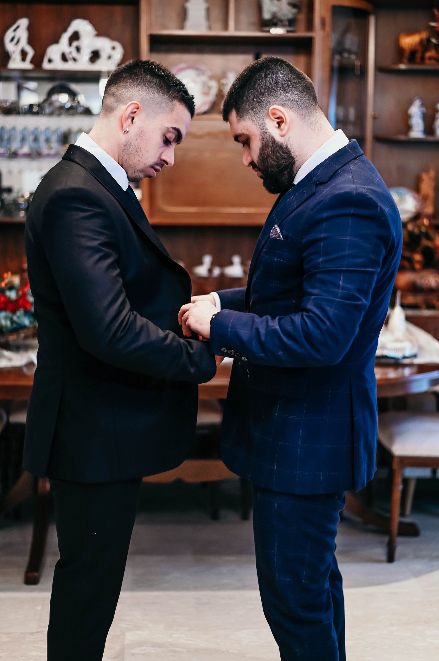 Two men in formal suits standing close together adjust cuffs and clothing details in an interior setting.