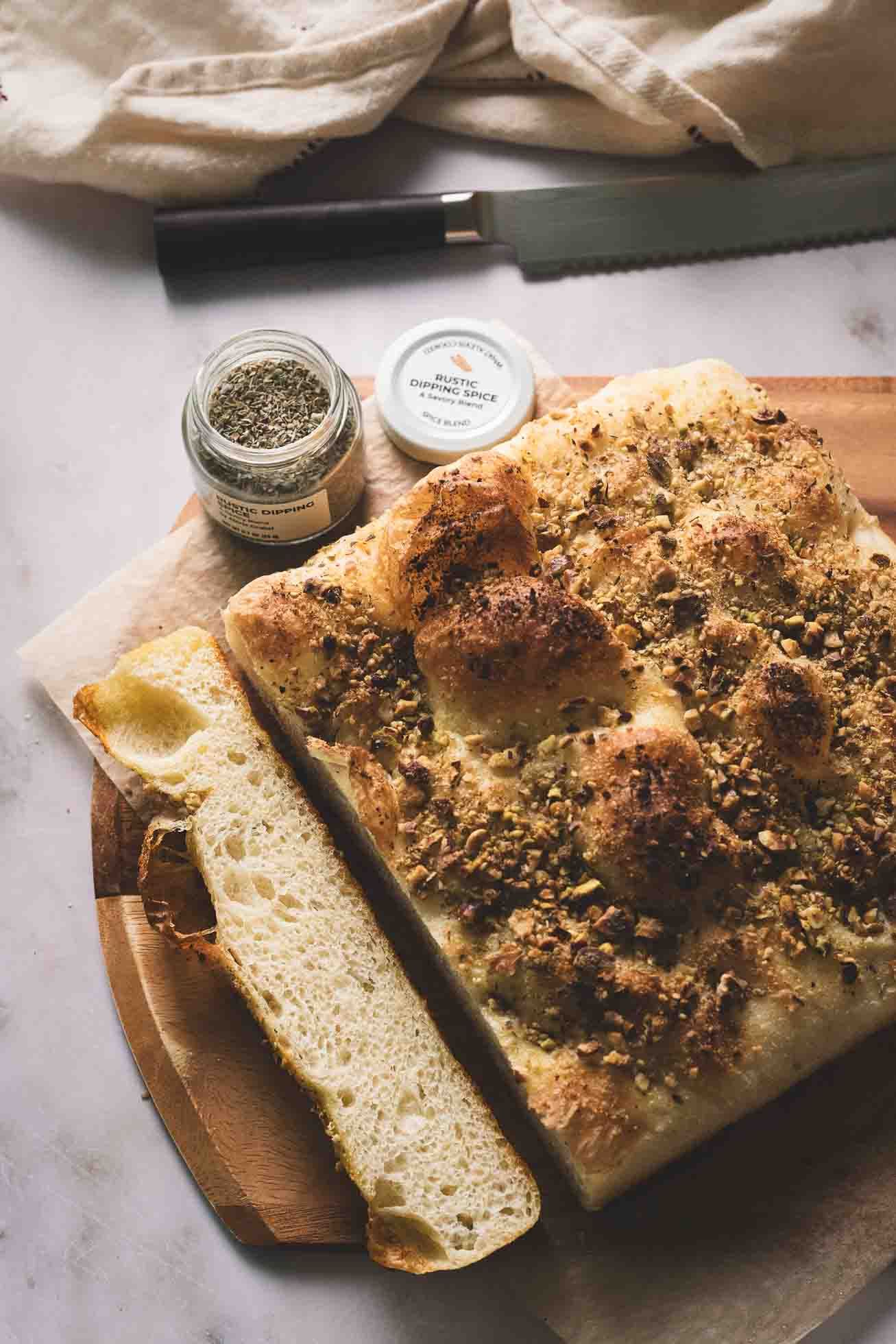 Freshly baked focaccia bread with herbs and seasonings on wooden board next to jar of spices and serrated knife.