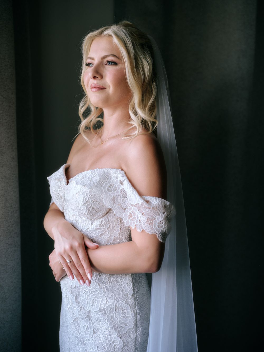 A bride in an off-shoulder lace wedding dress poses elegantly against a dark background. Photoshoot by Venice Photographer Alina Indi