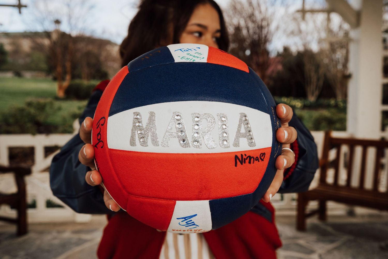 A red, white and blue volleyball with the word MAYA written on it is held up against an outdoor background.