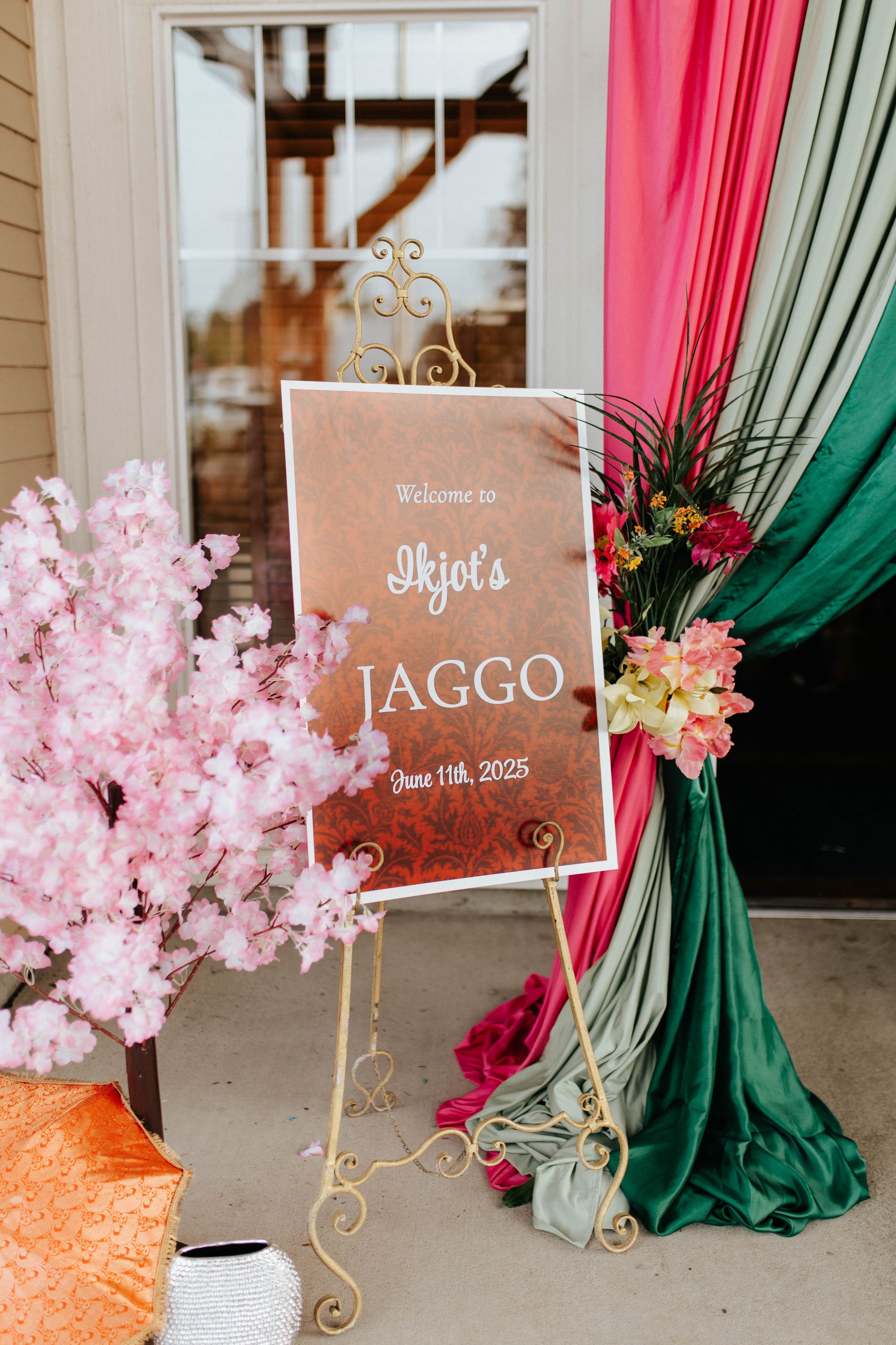 Welcome sign on ornate gold stand with pink cherry blossoms and red and green draped fabric at an elegant Jaggo event.