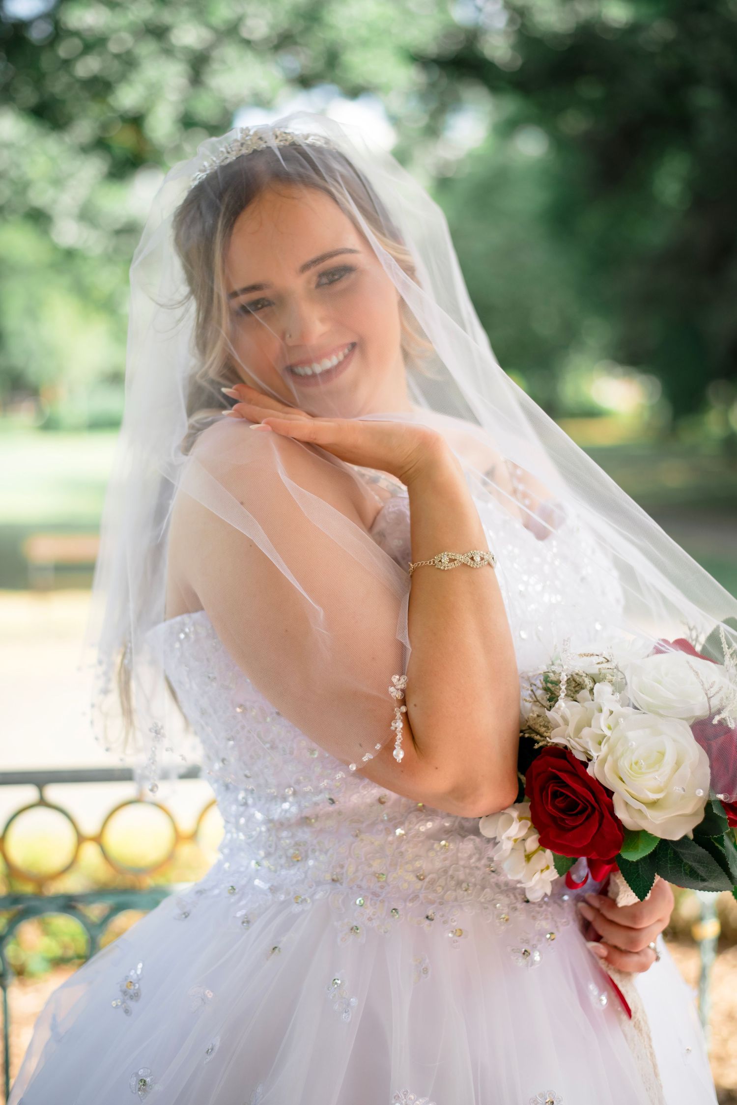A bride in a white beaded wedding dress holding a red and white rose bouquet smiles during her wedding celebration.