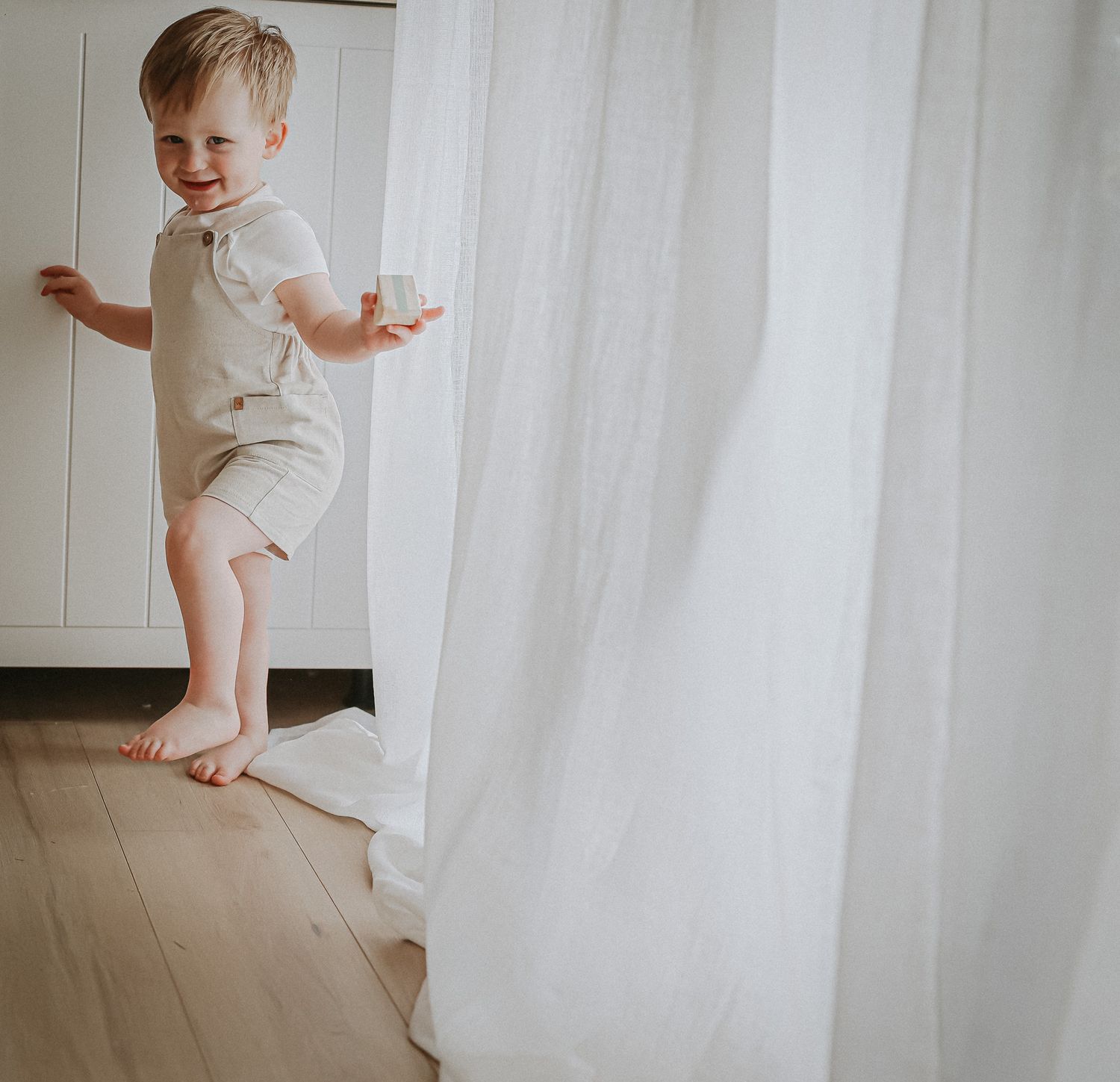 A young child in a white outfit plays near sheer curtains on a hardwood floor.